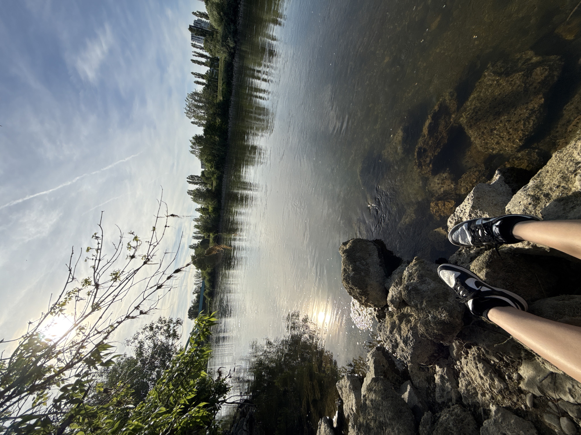 The photograph shows a view of a riverbank in Zagreb, Croatia, on a sunny day. The foreground is dominated by rocky terrain where a person sits, with their legs and sneakers visible. The river flows from the center to the upper right, reflecting the sunlight and trees along its banks. In the background, buildings and trees line the opposite shore. The sky is partly cloudy with contrails. Vegetation, including trees and shrubs, is present on the near bank.
