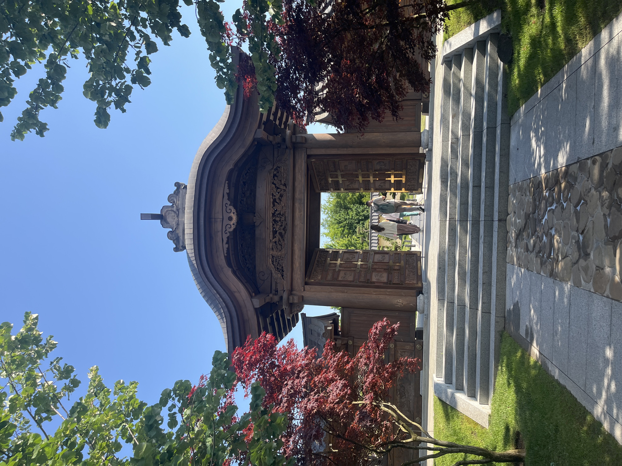 Two individuals walk through an open wooden gate, framed by trees and a clear blue sky. The gate is ornately carved and appears to be part of a traditional, possibly Asian, architectural style. Stone steps lead up to the gate, and a paved path with rounded stones runs in the foreground. The scene is bathed in sunlight, casting shadows. This image depicts the entrance to the Krasnodar Buddhist Temple, located in Krasnodar, Russia.