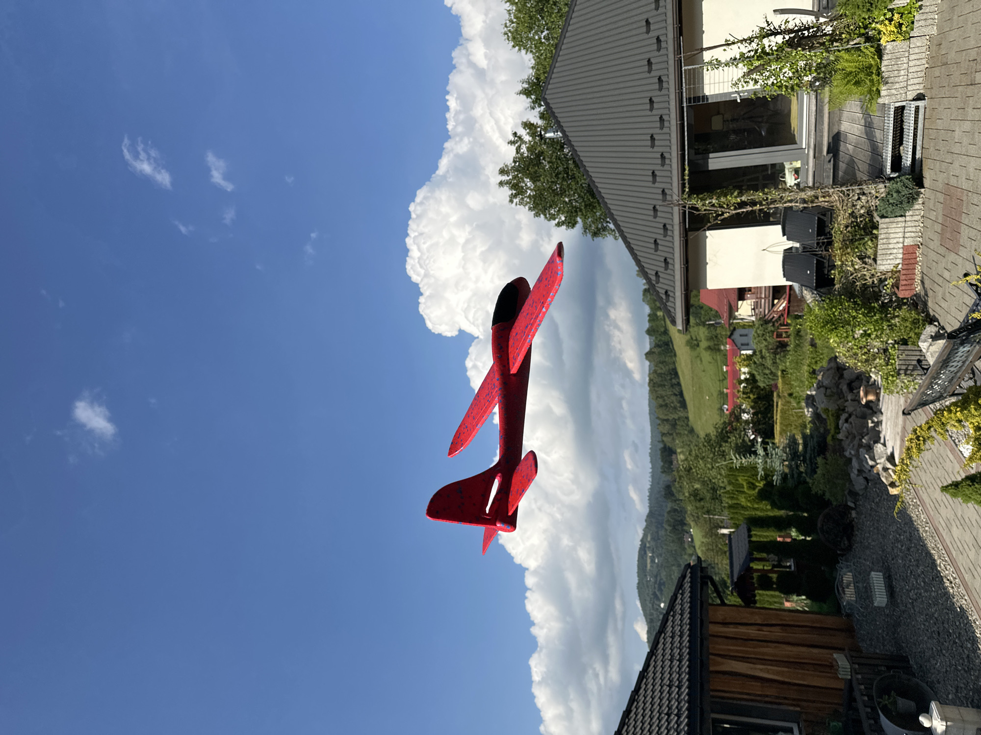 The image depicts a bright, sunny day in a suburban or rural setting, likely in or near Wisła, Poland, characterized by lush greenery and rolling hills in the background. A bright red toy airplane with blue speckles is prominently featured in the foreground, appearing to be in mid-flight against a clear blue sky dotted with fluffy white clouds. In the middle ground, the rooflines of several houses are visible, one with a dark, corrugated metal roof and wooden siding, and another with a white facade and dark-trimmed windows. The surrounding landscape is filled with mature trees and various types of foliage, including manicured bushes and garden plants. The foreground shows a paved patio area with stone accents and garden beds. The overall impression is one of a peaceful, idyllic outdoor scene.