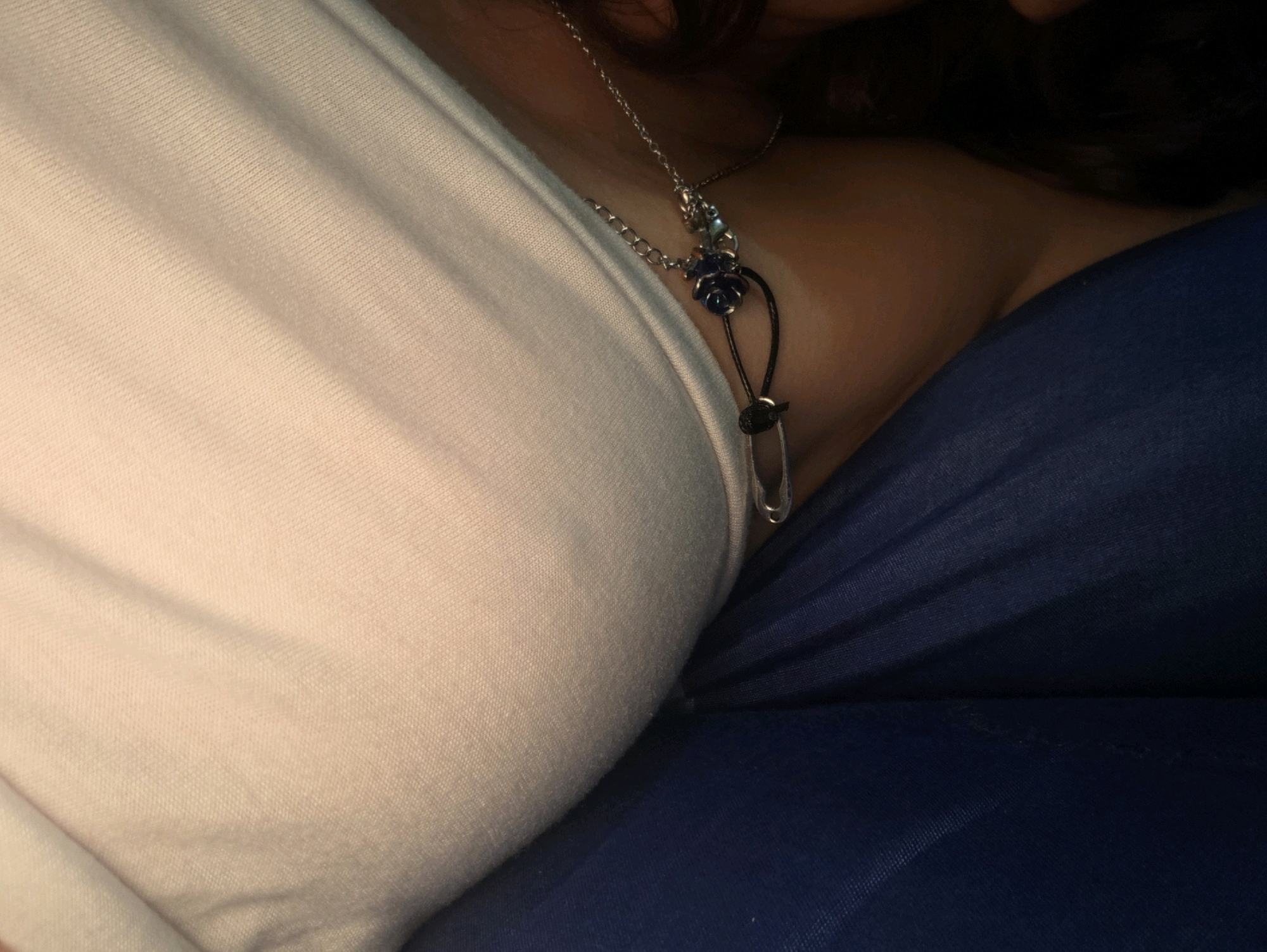The image is a close-up shot of a person's neck and upper chest area, draped with a light-colored fabric, possibly a t-shirt. A delicate silver necklace with a dark blue rose charm and a small safety pin pendant is visible. The person appears to be reclining, with a dark blue fabric in the background, suggesting they are sitting or lying down. The lighting is dim, creating shadows and highlighting the texture of the fabrics and the jewelry. There is no discernible urban or natural environment visible. The image does not contain information about the location in Phan Thiết, Vietnam.