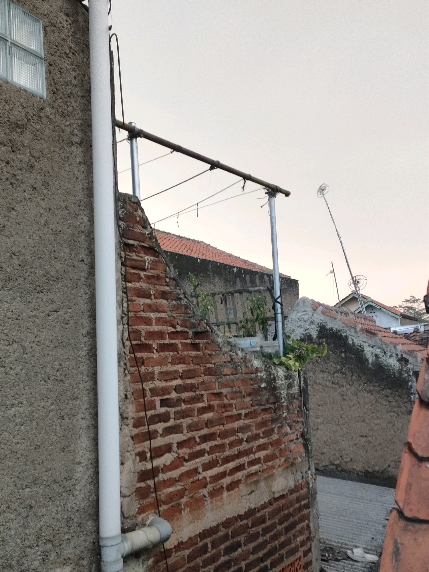 The image depicts an outdoor scene, likely in a residential area of Sumedang, Indonesia. In the foreground, a textured gray concrete wall runs vertically on the left, with a small glass block window near the top. A white PVC pipe runs down this wall, connected to a smaller gray pipe with an elbow fitting at its base. Adjacent to this wall is a section of weathered red brick wall, showing exposed mortar. A metal pole supports a horizontal bar with clothesline wires strung between it and another pole further back. Several other structures, including tiled roofs and concrete walls, are visible in the background, along with some sparse vegetation. A thin, long antenna-like structure is visible on the right side of the image. The sky is overcast and a pale white, suggesting either early morning or late afternoon. No people are present in the scene.