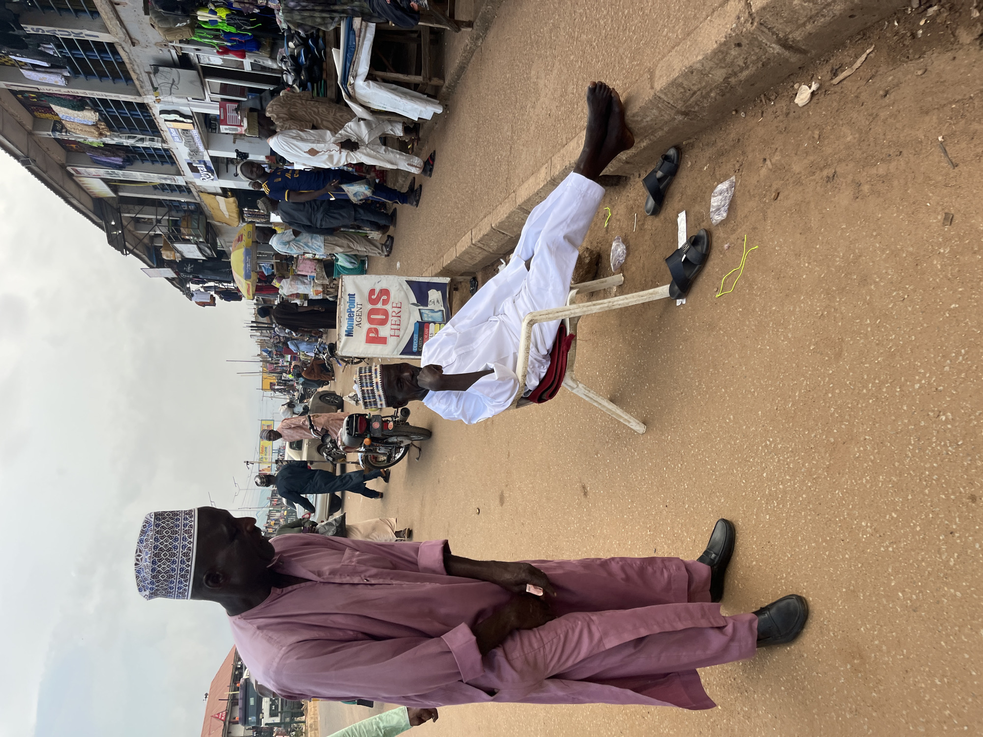 The image shows a street scene in Bukuru, Nigeria. In the foreground, two men are present. The man on the left is standing and wearing a pink traditional outfit and a patterned cap. He is holding a small white object in his hand. The man on the right is seated on a white folding chair, wearing a white traditional outfit and a cap. He is leaning back, resting his head on his hand. His feet are bare and resting on the curb. There are two pairs of black sandals on the ground near him. A yellow string is also visible on the ground. In the background, a busy marketplace is depicted with stalls selling various goods, including clothing. Several other people are walking through the market, and a motorcycle is parked. A sign for "MoniePoint AGENT POS HERE" is prominently displayed. The ground is a dusty, reddish-brown color. The sky is overcast.