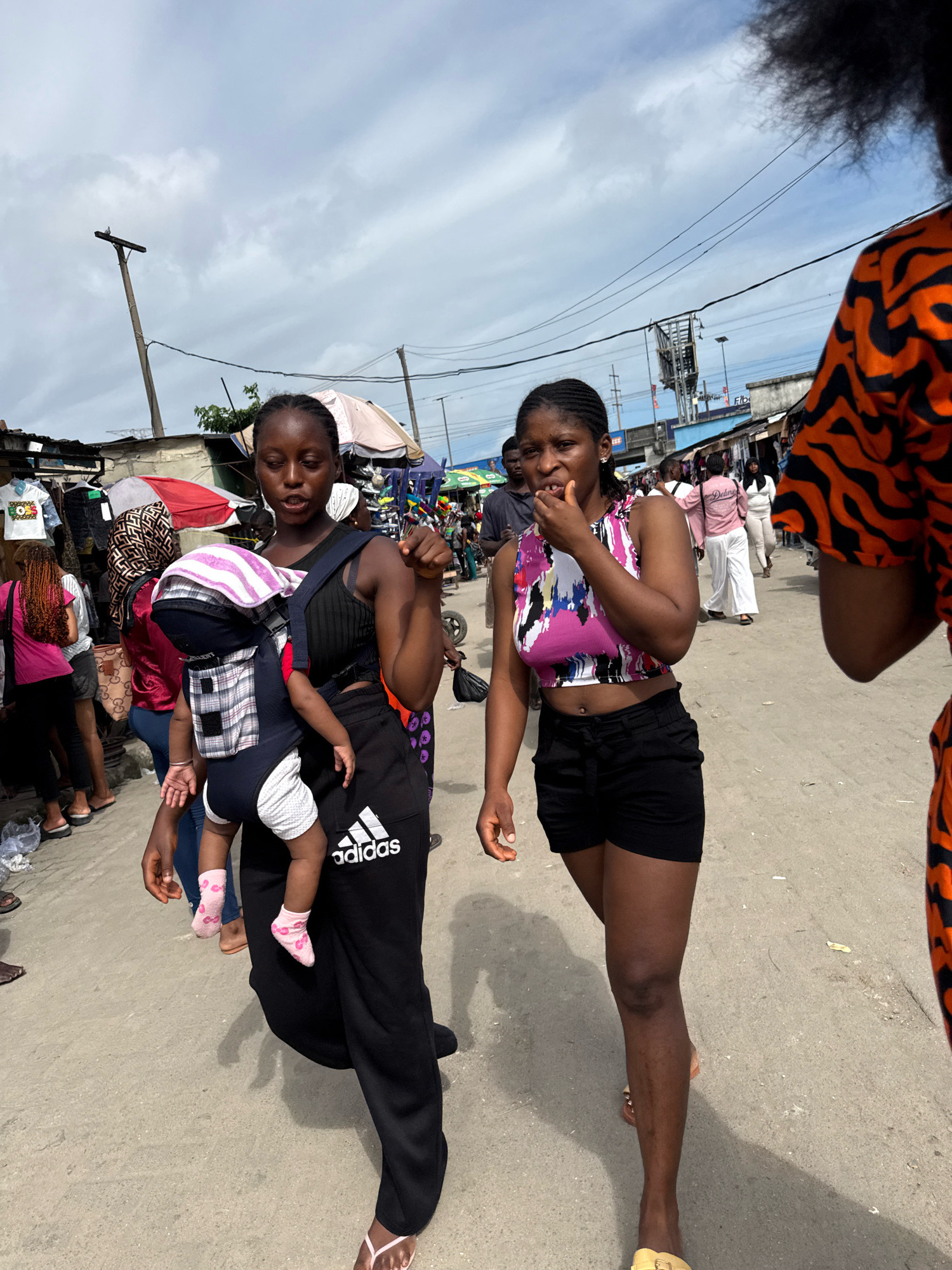 In a bustling marketplace in Lagos, Nigeria, two young women walk side-by-side. The woman on the left carries a baby in a front-facing carrier, the infant's legs dangling out, clad in pink socks. She wears a black tank top and black Adidas sweatpants. The woman on the right sports a colorful crop top and black shorts, with her hand near her mouth. Behind them, the marketplace unfolds with stalls displaying clothing and various goods under makeshift awnings. The ground is a dirt or asphalt path, and overhead, electrical wires crisscross under a cloudy sky. Other people can be seen in the background, adding to the lively atmosphere of the market.