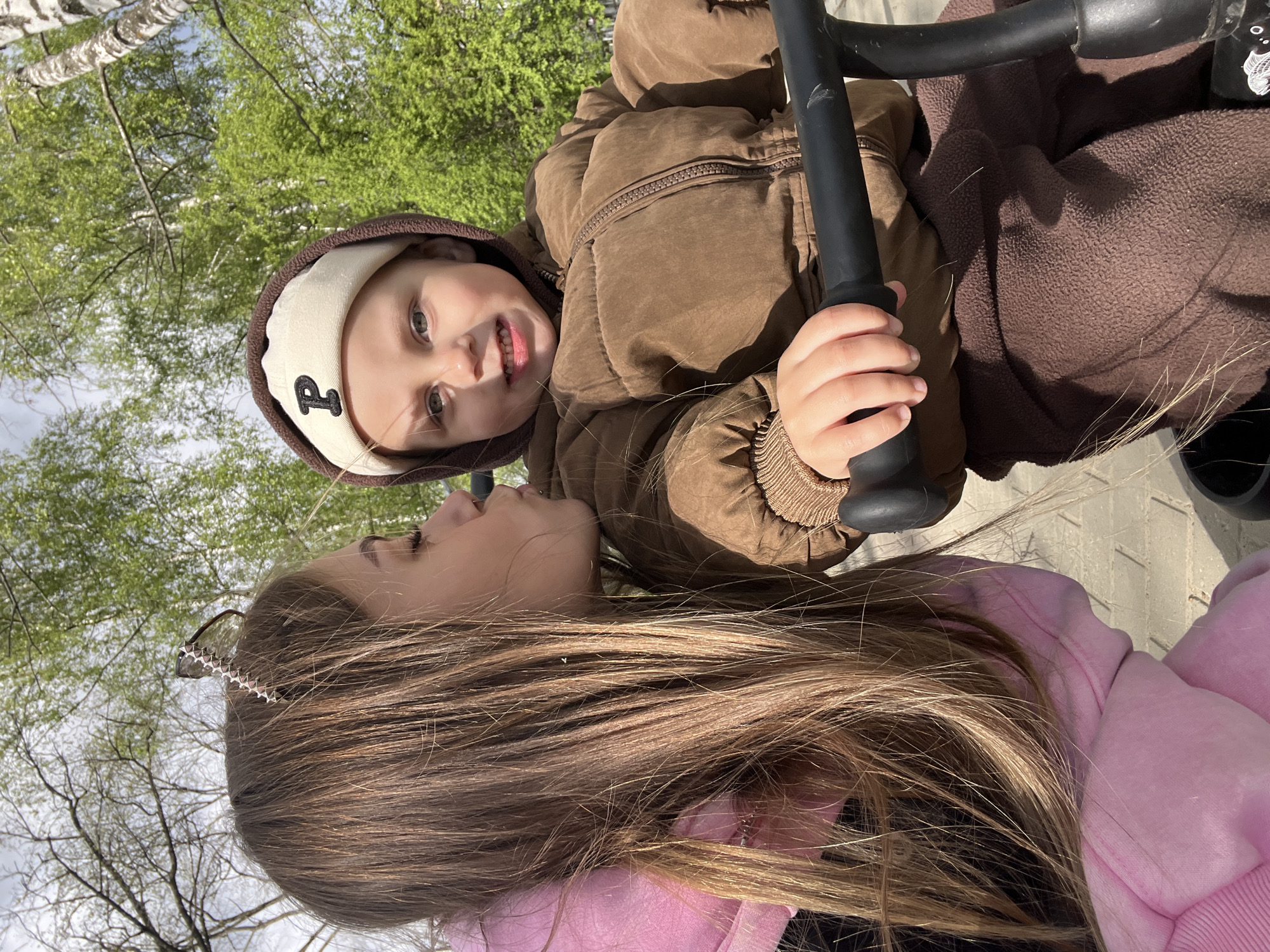 A tilted medium shot reveals a child in a brown hooded jacket holding onto a black bar, possibly part of a swing set. Beside the child, a person with long, brown hair wearing a pink hooded sweatshirt is visible. The setting appears to be outdoors, with trees and foliage in the background. Given the request context, the location might be in Chaikovsky, Russia.