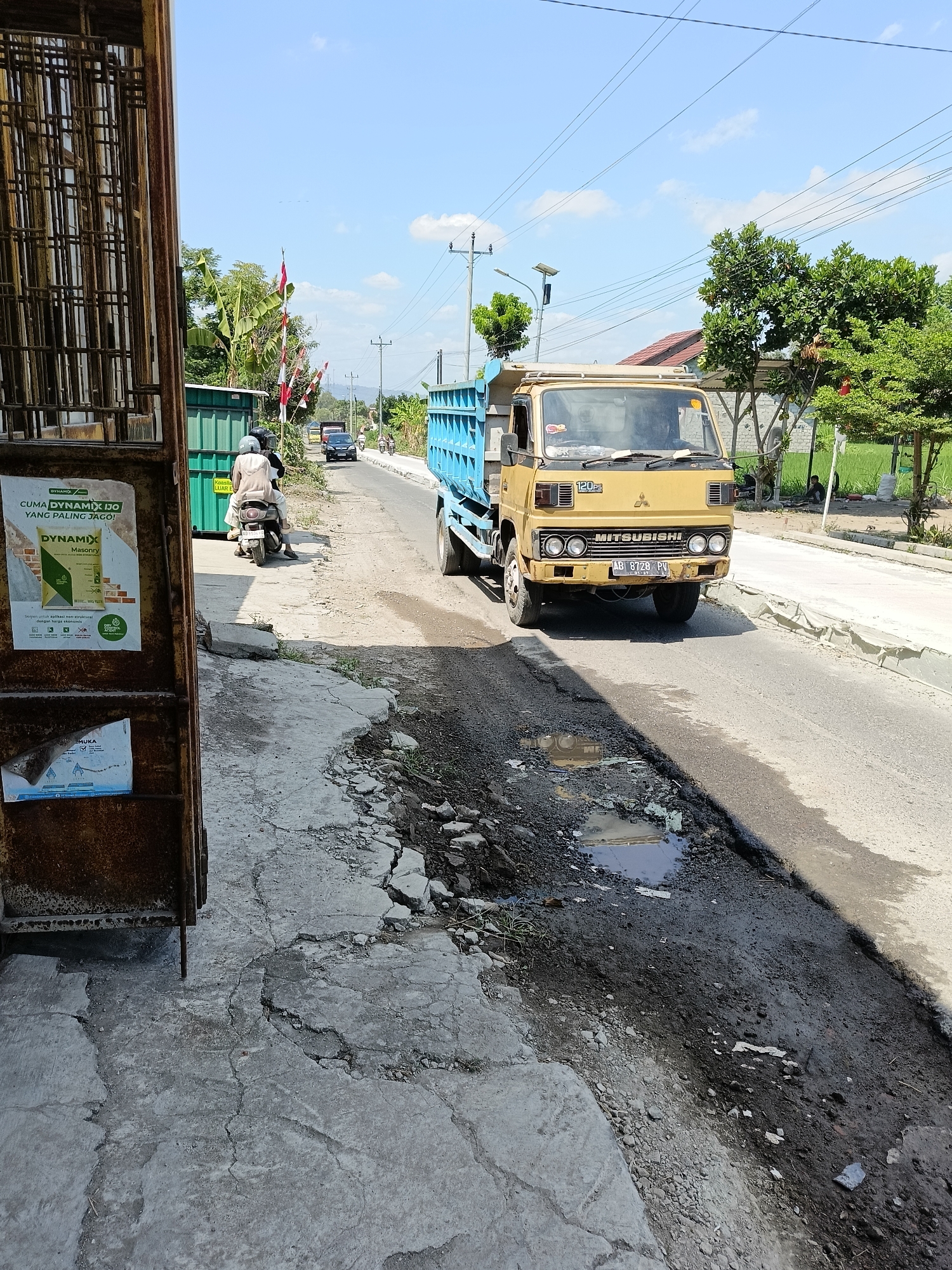 A yellow Mitsubishi dump truck is driving down a paved road. The truck has a blue bed. There is a motorcycle with two people on it in front of the truck. The road is lined with trees and greenery on the right side. On the left side of the road, there is a rusty metal gate with a poster on it. The road is cracked and uneven in some places, with puddles of water. The sky is blue with some clouds. The scene appears to be in a rural or suburban area of Sukorini, Indonesia, as indicated by the presence of Indonesian flags and vegetation.
