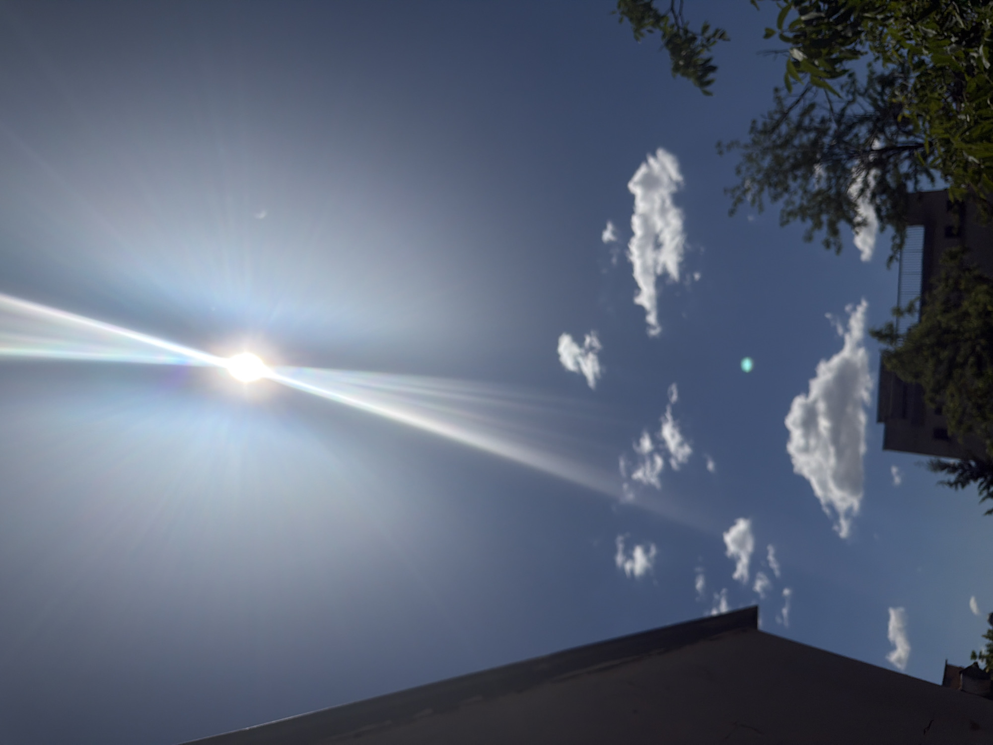 The image shows a daytime sky in Plovdiv, Bulgaria. The sun is a prominent feature, emitting bright light and rays. Several small, white clouds are scattered across the blue sky. A building with foliage is partially visible in the upper right corner. The edge of a rooftop is visible in the lower portion of the image.
