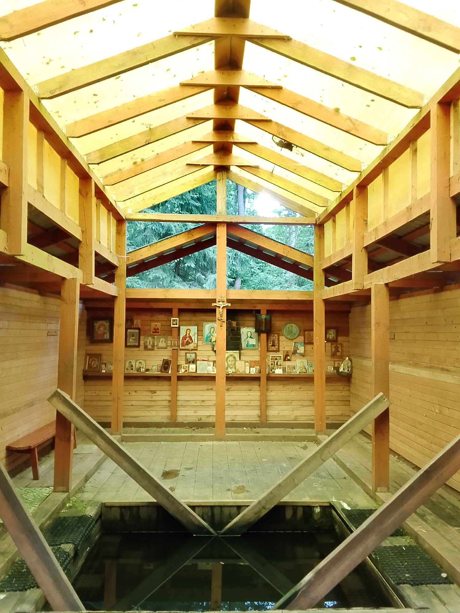 The image depicts the interior of a wooden structure, likely a chapel or a baptismal site, in Staryi Pokrov, Russia. The structure features a sloped roof made of yellow translucent material supported by wooden beams, allowing diffused natural light to filter in. The walls are constructed of horizontal wooden planks, and shelves are mounted on them, displaying numerous religious icons in frames of various sizes. A cross with a figure of Christ is visible on the right wall.
In the center of the structure, there is a dark pool of water, possibly a holy spring or a baptismal font, with wooden beams angled over it, serving as a decorative element or a guide. Wooden walkways surround the pool. To the left, a simple wooden bench is placed against the wall. The background reveals a view of dense green trees through an open section of the structure, suggesting an outdoor, possibly forested, location. The overall atmosphere is serene and reverent, with the warm tones of the wood and the diffused light creating a tranquil ambiance. The time of day appears to be daytime, with overcast or cloudy weather suggested by the diffused lighting. No visible text is present in the image.
