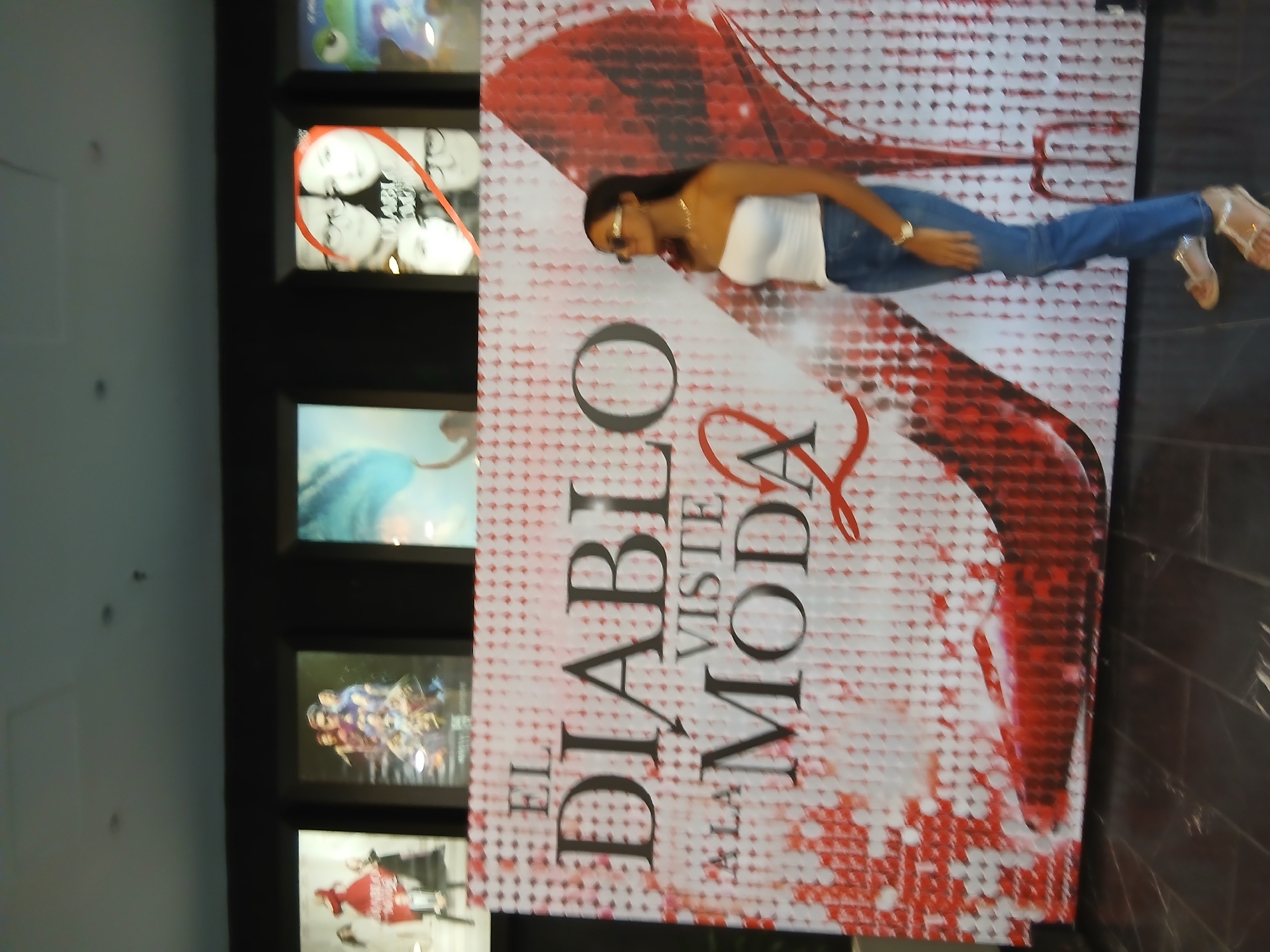 The image shows a woman posing next to a movie poster. The poster reads "El Diablo Viste A La Moda." The woman is wearing a white top, jeans, and sandals. She is wearing sunglasses. The scene appears to be indoors, next to a wall with several movie posters. The specific location in the city of Altagracia, Venezuela, is not determined from the image.