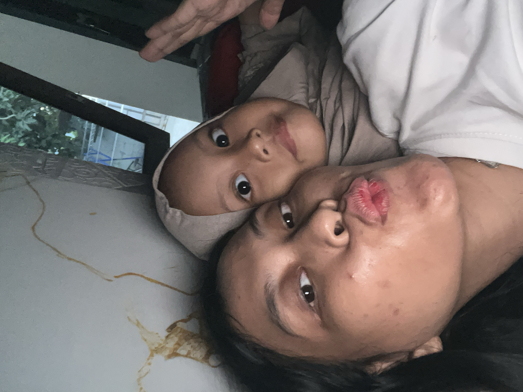This is a close-up, vertically oriented, selfie-style photo of two people, likely a mother and child, lying down. The subjects are framed in a way that their faces are the primary focus, with the child's face positioned above and slightly to the left of the adult's. The child is wearing a light brown hijab, and their eyes are wide open, looking directly at the camera. The adult is wearing a white t-shirt and is leaning in towards the child, puckering their lips as if for a kiss. Their eyes are also open and looking towards the camera.
The background is dark and out of focus, but appears to be an indoor setting with some foliage visible through a window or opening to the left. There are some brown, stain-like marks on a light-colored surface in the foreground, which could be a mattress or a table. The lighting suggests it might be daytime, but the exact time is not discernible. The overall mood of the image is affectionate and playful, captured in a candid moment between the two individuals. There is no visible text in the image.