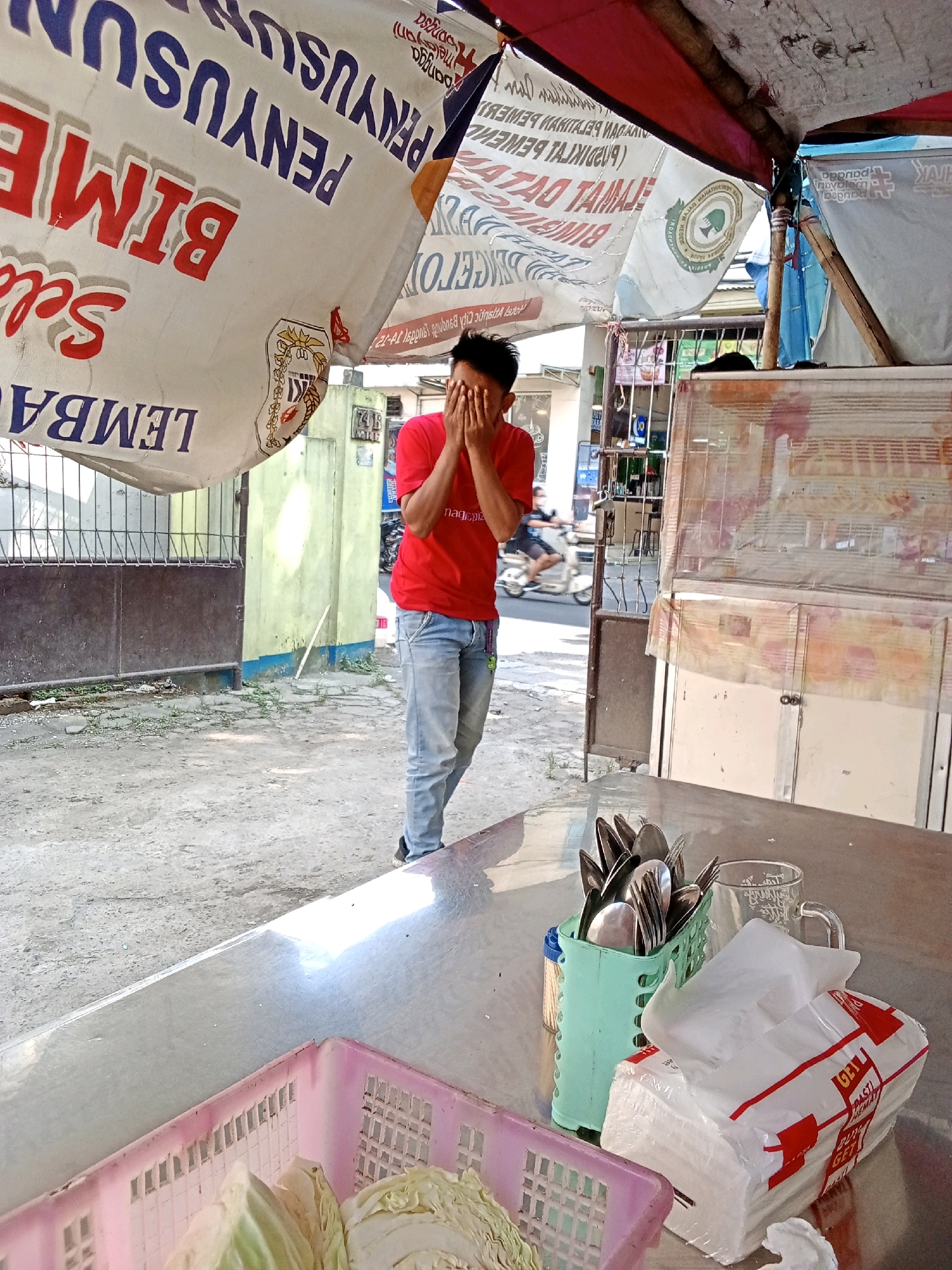 A young man wearing a red t-shirt and blue jeans is standing with his hands over his face, suggesting he is upset or hiding his emotions. He is outdoors in what appears to be a street market or food stall area. In the foreground, a stainless steel table is set with cutlery in a green holder, a glass, and a packet of tissues. A pink basket with cabbage is also visible on the table. Behind the man, a stall awning with Indonesian writing is prominent. The background shows a street with some buildings and a person riding a motorcycle. The environment appears to be a busy urban setting in Bandung City, Indonesia.