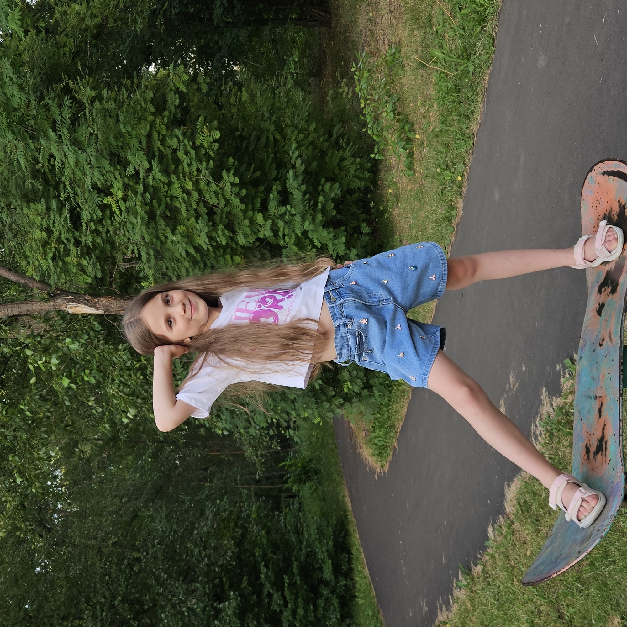 A young girl with long, blonde hair and a white t-shirt with pink lettering is posing on a skateboard. She is wearing blue denim shorts adorned with various small charms, possibly sea creatures and other figures. Her right hand is resting on her head, and she has a gentle smile on her face, conveying a sense of happiness and playfulness. She is wearing pink sandals.
The setting is outdoors, with a paved path winding through a grassy area. Lush green trees form a dense backdrop, suggesting a park or a wooded area. The lighting indicates it is daytime, likely a sunny day given the bright illumination. The skateboard is colorful, with patches of orange and blue, showing signs of wear. The girl's stance on the skateboard, with one leg extended, suggests she is either posing for a picture or about to ride it.
The visible text on her t-shirt appears to be "SURF" and "ESTINATIONS ONLY". The overall impression is of a candid moment capturing a child's enjoyment of a summer day and an activity. The context of Kemerovo, Russia, places the scene in a specific geographical location.
