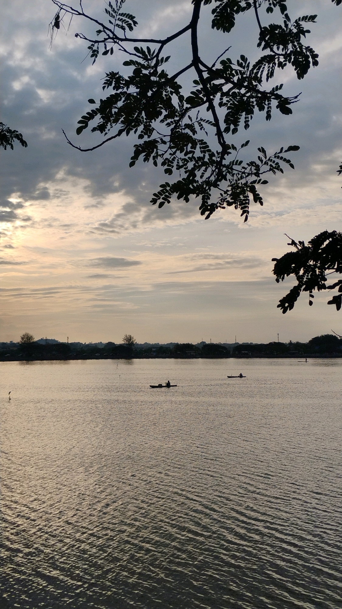 This image depicts a serene waterscape at dusk, likely in Lhokseumawe, Indonesia. In the foreground, silhouetted tree branches with small leaves frame the scene from the top. The middle ground is dominated by a calm body of water, reflecting the soft, diffused light of the setting sun. Two small boats, each with a solitary figure, are visible on the water. One boat is closer to the left bank, and the other is further to the right. A thin, dark silhouette of a bird is standing on a small pole in the water on the left side. The background features a low-lying, silhouetted shoreline with scattered trees and structures, suggesting a town or city in the distance. The sky is a gradient of muted colors, with soft clouds and the pale glow of the sun on the horizon, indicating the end of the day. The overall impression is one of tranquility and natural beauty.
