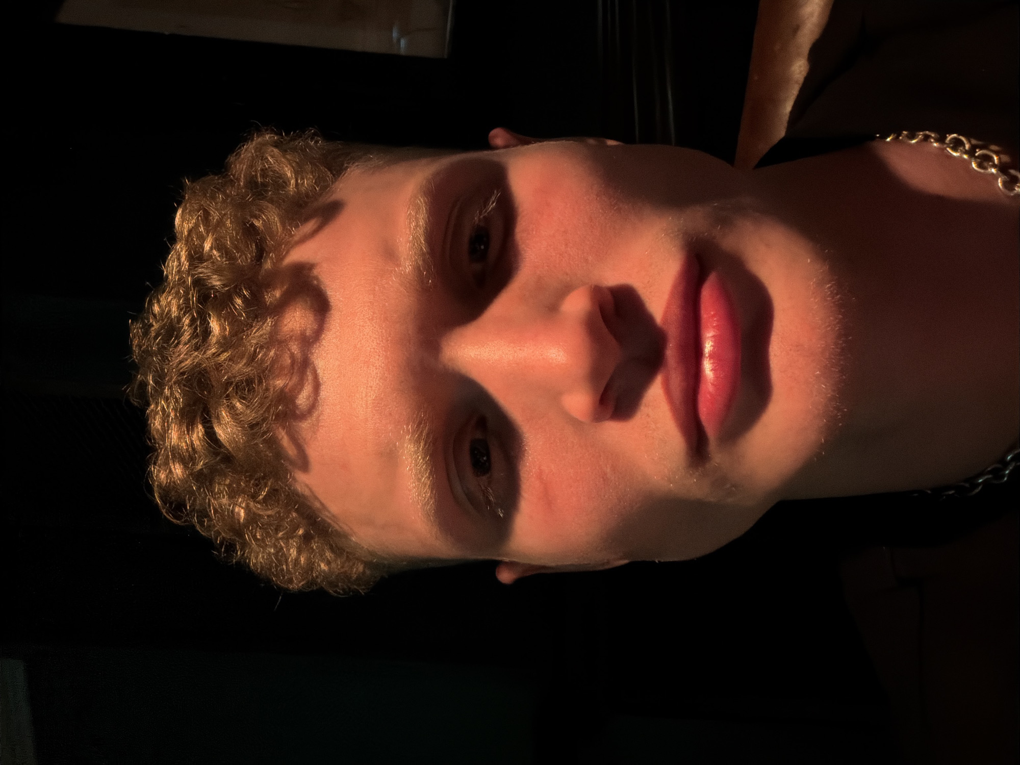 A close-up, high-contrast portrait shot captures the face of a person, likely male, with curly, light brown hair. The image is dramatically lit, with a warm, reddish-orange light source illuminating the left side of the face (when the image is viewed upright). This lighting highlights the cheekbone, nose, and lips, casting deep shadows across the right side of the face and the entirely dark background.
The subject is looking directly at the camera with a neutral expression. Notable features include prominent, full lips that appear glossy, a well-defined nose, and visible eyelashes and eyebrows. A metallic chain, likely silver or gold-toned, is visible around the neck or shoulder area on the illuminated side of the body. The background is completely obscured by shadow, offering no specific location cues beyond the dark, ambiguous setting. The overall aesthetic is one of intense and stylized portraiture, emphasizing contours and textures through the interplay of light and shadow. No visible text is present in the image.
