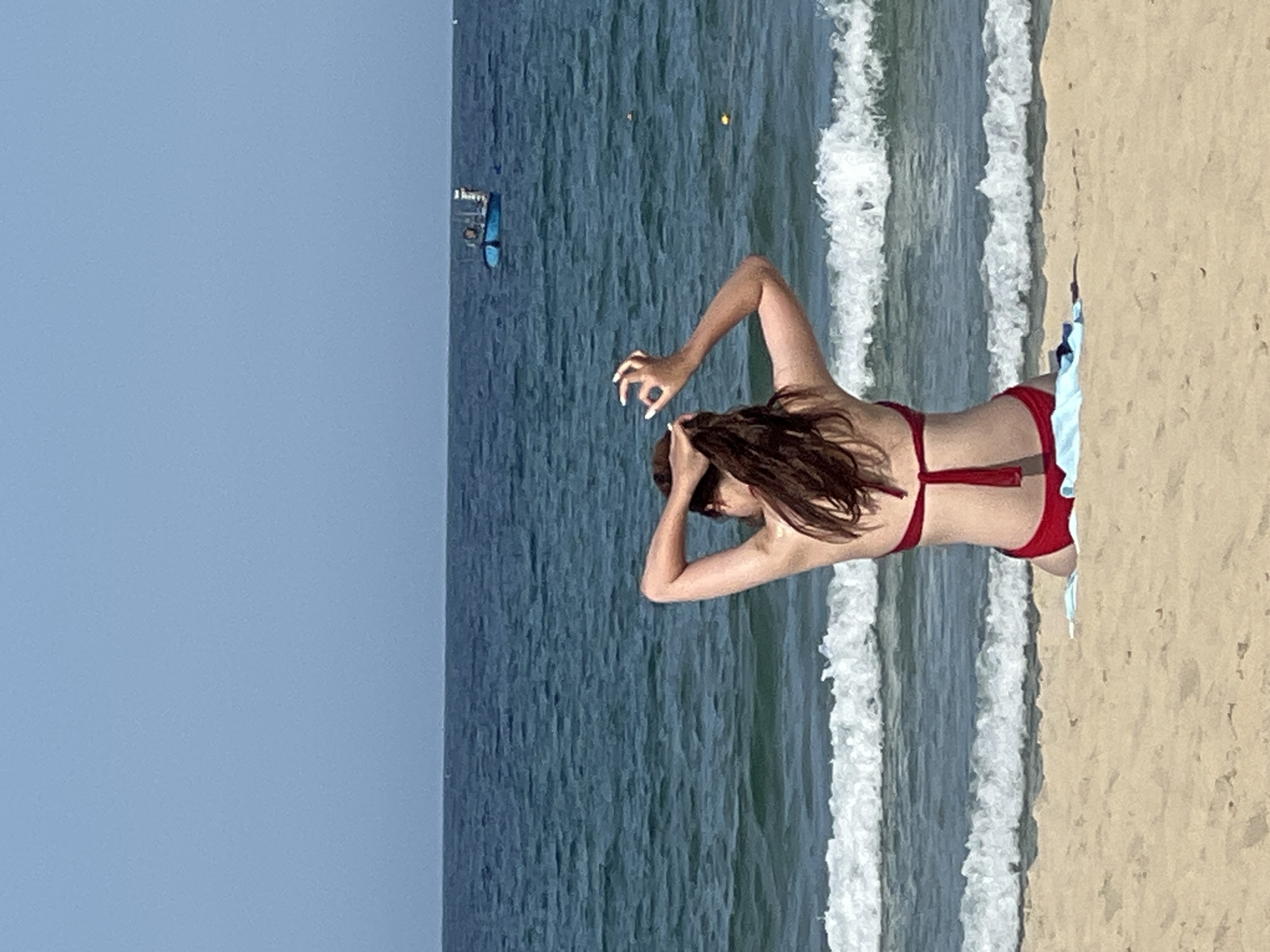 A woman in a red bikini is sitting on a sandy beach, facing the ocean. Her back is to the camera as she ties her dark brown hair up. Her left arm is raised, and her hand is positioned near her head, with her fingers splayed out in a gesture. The ocean is blue with white-capped waves, and the sky is a clear, pale blue. In the distance, a small blue boat is visible on the water. The scene takes place on a beach in Đà Nẵng, Vietnam.