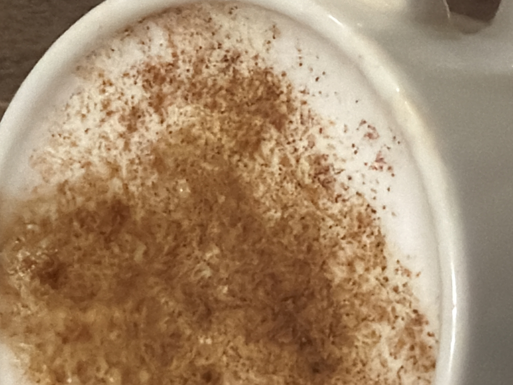 The image shows a close-up of a white cup filled with a beverage, likely a latte or cappuccino, topped with a layer of foamed milk and sprinkled with a brown spice, possibly cinnamon or nutmeg. The cup appears to be sitting on a wooden surface. The setting is unknown.