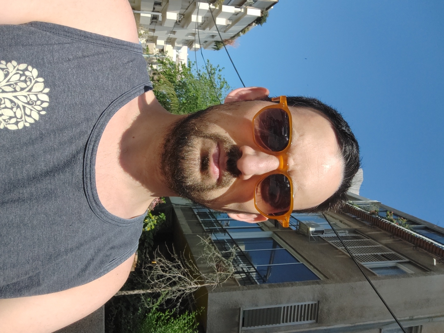 A medium shot captures a man wearing a grey tank top adorned with a white tree-like design on the upper left chest. He sports dark, slightly overgrown hair, a well-trimmed dark beard, and sunglasses with orange frames and dark lenses. He is looking directly into the camera.
The background reveals an urban setting, with trees and greenery visible in the lower portion. Above, there are light-colored buildings, typical of Tel-Aviv architecture, featuring windows and balconies. A clear blue sky fills the upper frame, suggesting a sunny day. The overall composition indicates an outdoor setting in Tel-Aviv, Israel.