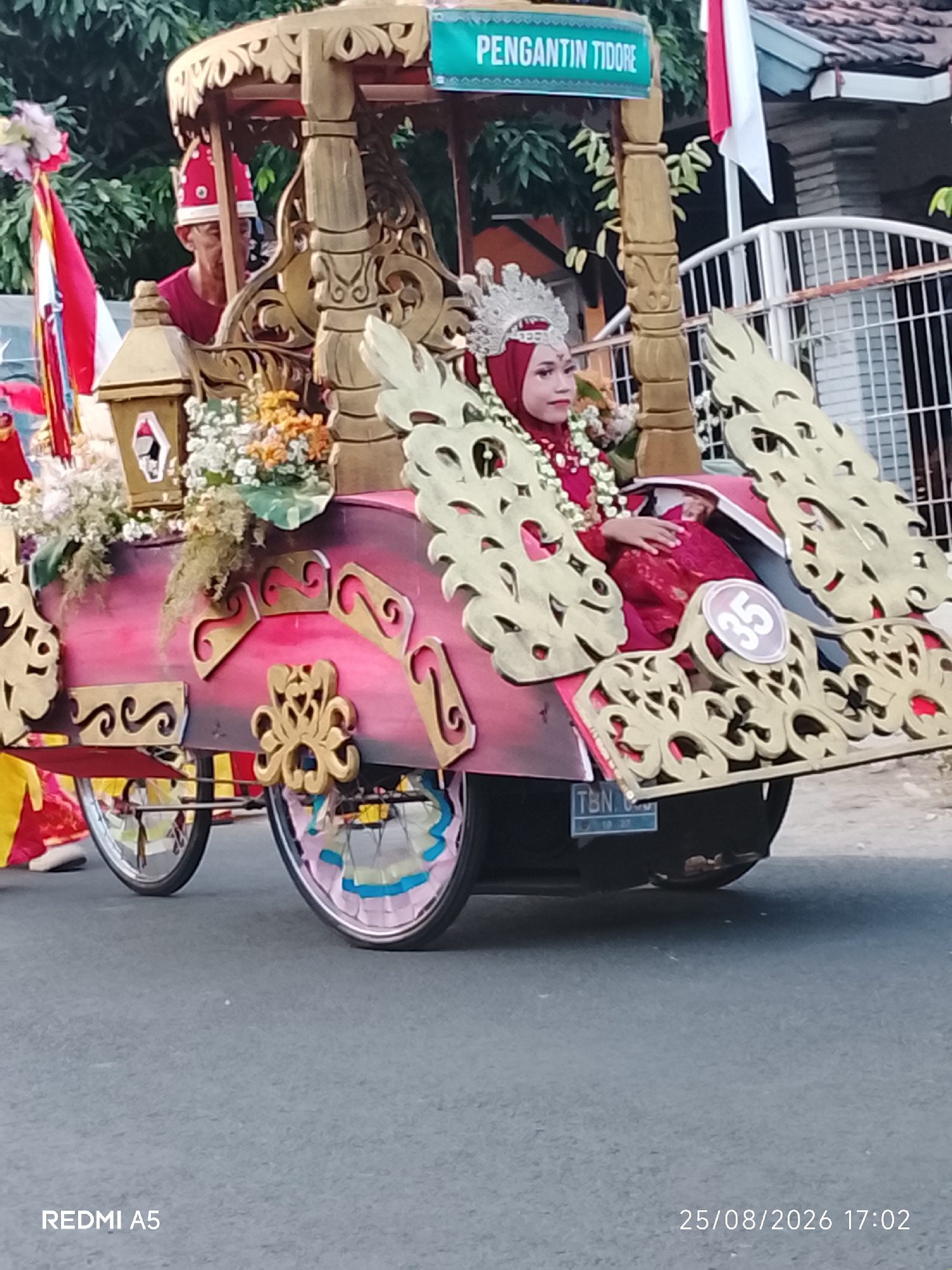 In Semanding, Indonesia, a woman in a red hijab and traditional headdress sits in an ornate, pink and gold carriage. The carriage is decorated with gold filigree and flowers, and it is pulled by unseen means. She is wearing a red dress and her face is painted with makeup. The carriage has wheels, one of which is decorated with colorful bands. A sign on the carriage reads "PENGANTIN TIDORE". A man in a red hat is partially visible behind her, and a red and white flag is displayed on the carriage. A license plate that reads "TBN 10 27" is visible on the back of the carriage. The scene appears to be a parade or procession, with a paved road and trees in the background. The image is dated 25/08/2026 17:02.