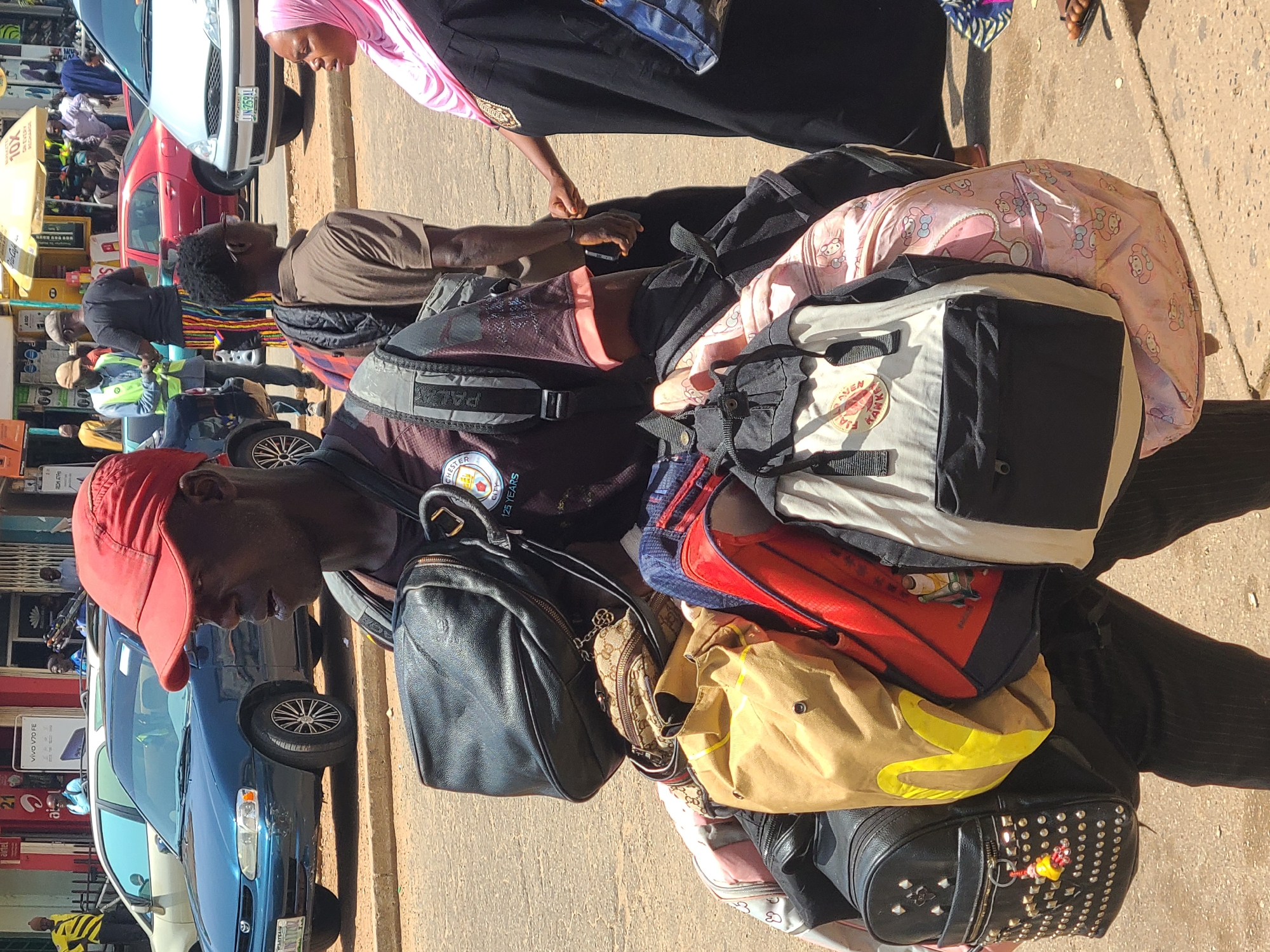 In Jos, Nigeria, a Black man wearing a red baseball cap and a dark t-shirt is walking, heavily laden with multiple bags. The bags include a black leather backpack with metal studs, a tan canvas bag with a yellow logo, a red bag, and several other backpacks and duffel bags, some with colorful patterns. He is also carrying a black shoulder bag. The man is in the middle of a street, with cars parked and driving in the background. A blue sedan is on the left, and a silver sedan is on the right. Other pedestrians are visible in the background, including a woman wearing a pink hijab. The environment appears to be a busy urban area with shops and businesses lining the street. The lighting suggests it is daytime, with shadows indicating sunlight. The ground is paved with asphalt and concrete.