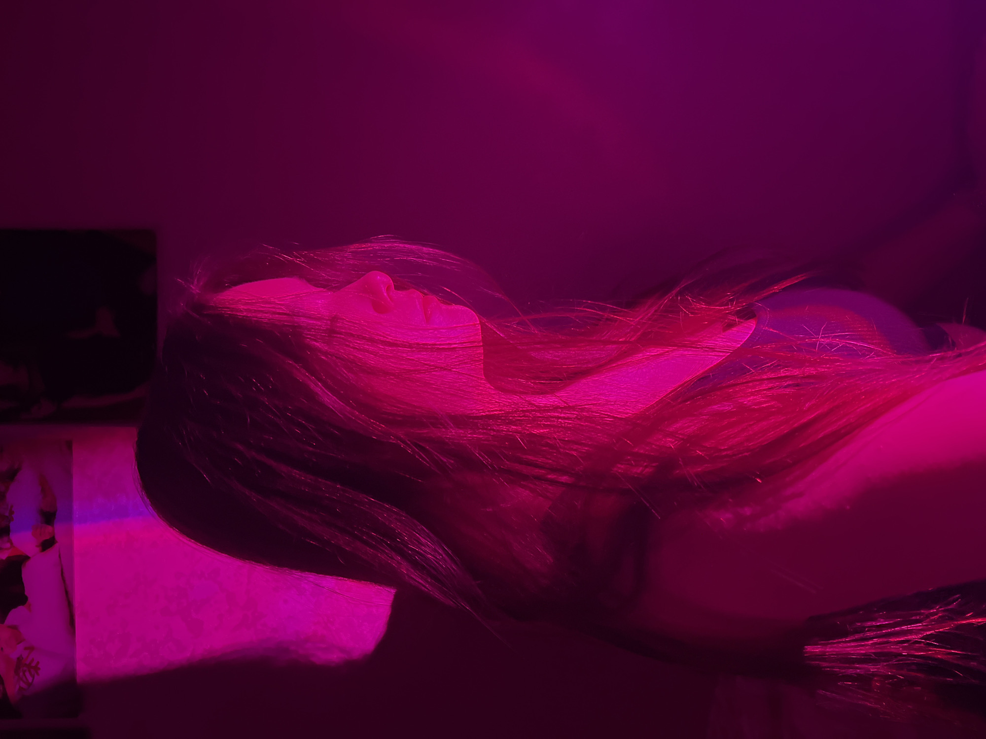 A person with long hair is lying on a pillow. The scene is illuminated by pink light. Part of a picture or poster is visible on the wall. The location is unknown, and there is no indication of it being in Chaikovsky, Russia.