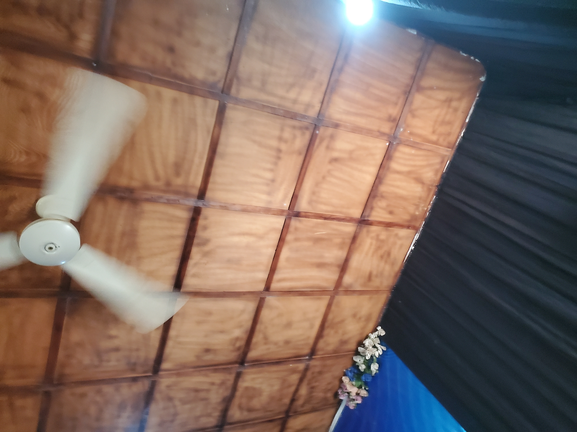 The image depicts a ceiling fan with its blades blurred due to motion, indicating it is in operation. The ceiling itself is constructed from wooden panels with visible dark brown dividing strips, forming a grid pattern. In the lower left portion of the image, a cluster of artificial flowers in shades of pink, purple, and white is visible. Beneath the ceiling and flowers, a blue surface transitions into a black, draped fabric, suggesting an indoor setting. A bright light source is visible in the upper right corner, possibly a ceiling light. The scene is located in Oturkpo, Nigeria.