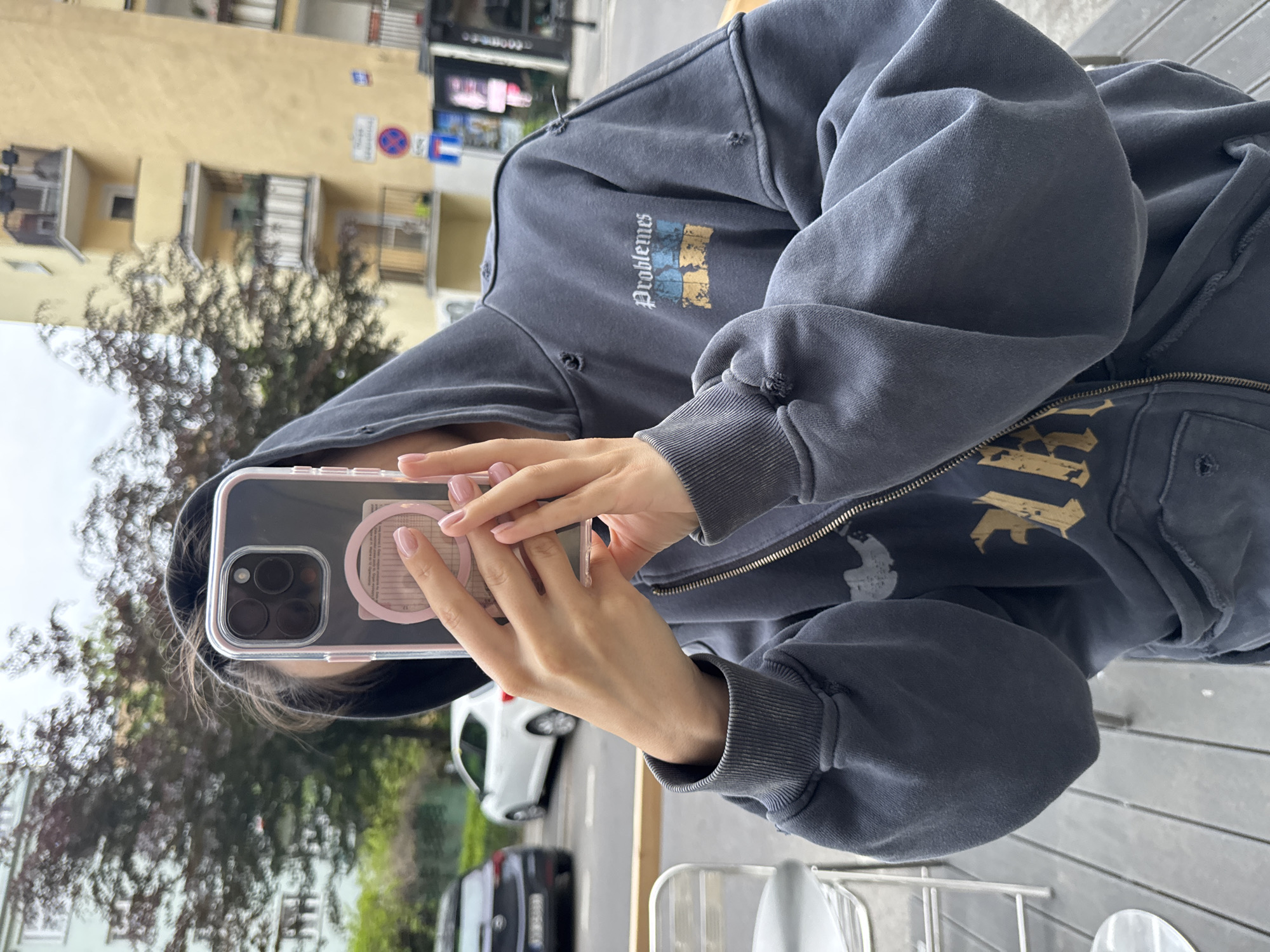 A medium shot shows a person in a hooded sweatshirt taking a photograph or video using a smartphone. The person is positioned on what appears to be a cafe or restaurant patio with a gray wooden table in the foreground, identifiable by the aluminum frame of a chair. The person is wearing a dark gray hooded sweatshirt with graphic prints in yellow, blue, and white. The phone has a transparent case with a pink circular grip attached to the back. A building with beige walls, visible signs and balconies appears in the background, with greenery of trees. Cars park along a street.