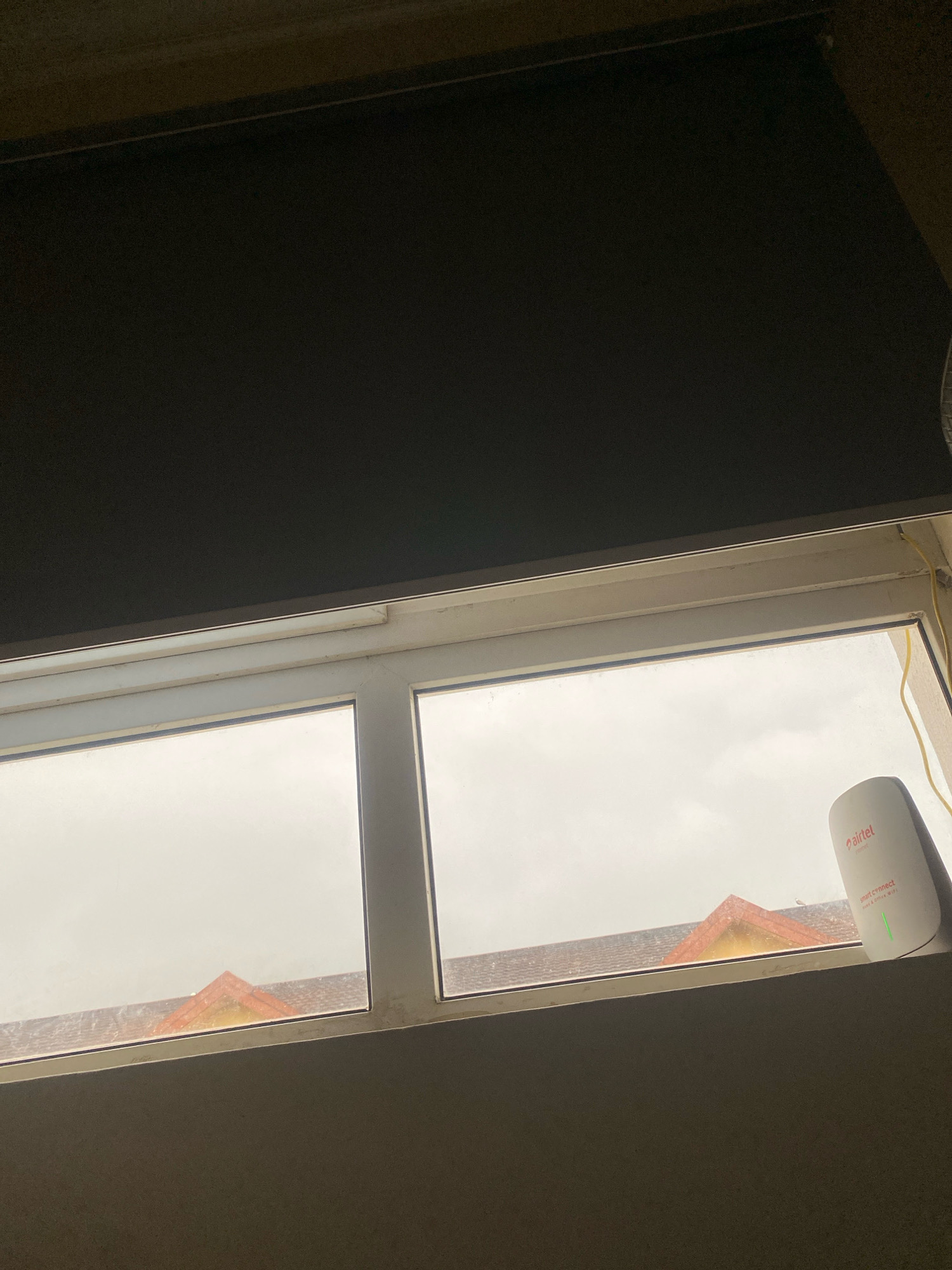 A window, partially obscured by a dark roller blind, reveals a view of rooftops under an overcast sky. In the foreground, a white Airtel "Smart Connect" router with a green indicator light sits on the windowsill. The window is divided into two panes by a vertical frame. The rooftops visible are clad in dark tiles, with a prominent orange-tiled peak visible on the right side. The overall scene suggests an indoor perspective looking out onto a residential or commercial area in Lekki Peninsula II, Nigeria.