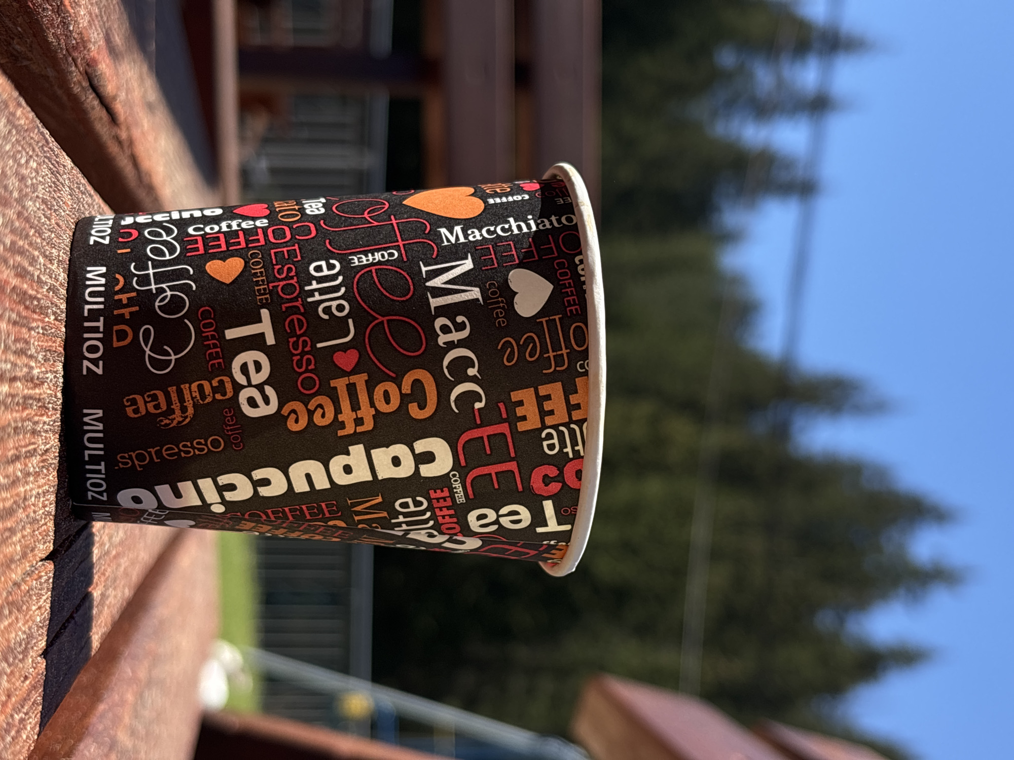 A close-up shot captures a coffee cup resting on a wooden surface, likely a table or bench. The cup is adorned with coffee-related words such as "Cappuccino," "Macchiato," "Coffee," "Tea," and "Espresso" printed in varying fonts and sizes. The backdrop features a blurred view of greenery, suggesting trees or foliage, and a clear blue sky, indicating a daytime scene. The location is unspecified.