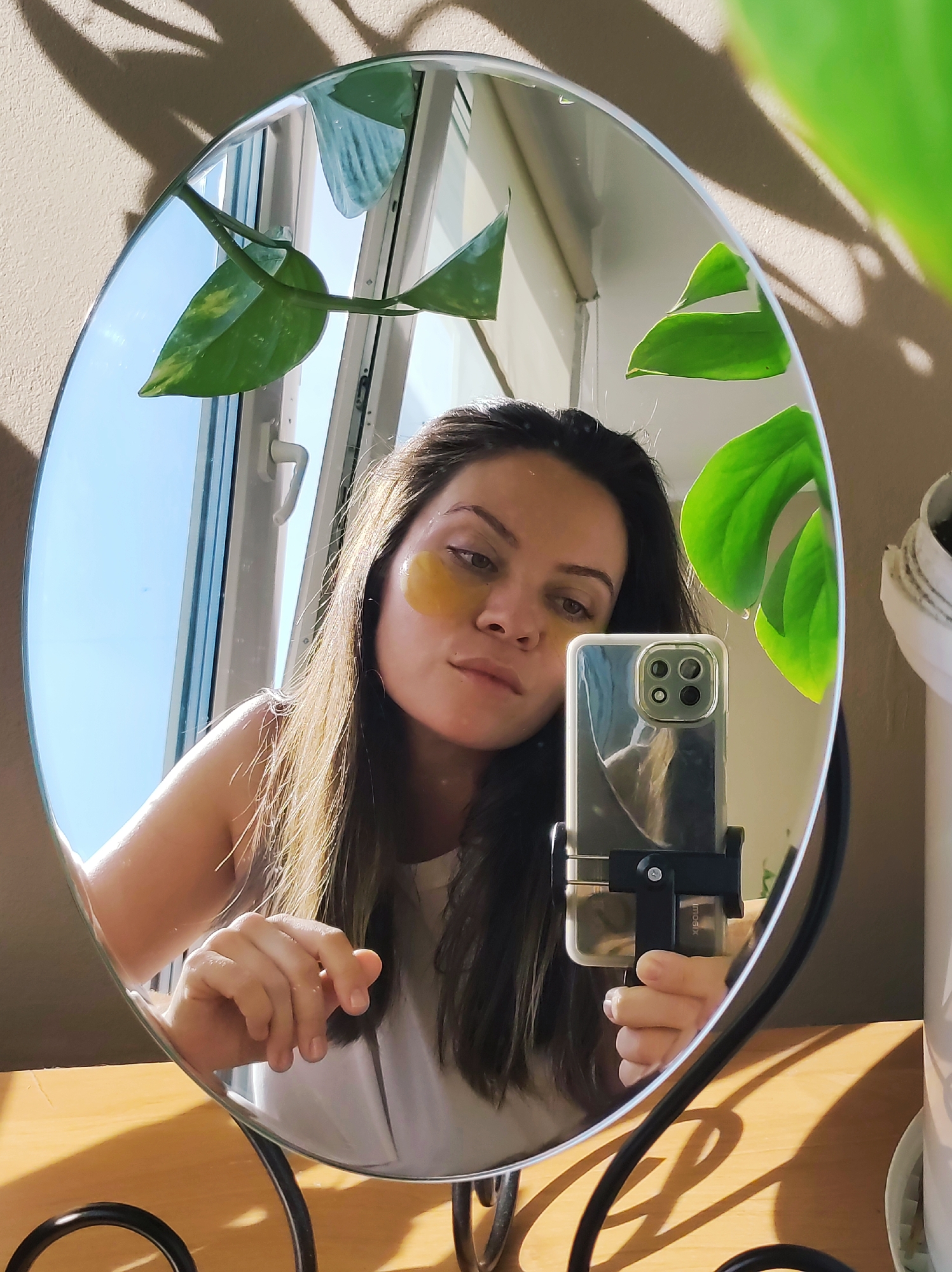 A mirror selfie shows a woman with dark hair, wearing gold under-eye patches. She is holding a smartphone in a mount, taking the photo in the mirror. The reflection includes a window with a clear sky and plants, both real and reflected. The mirror is standing on a wooden surface. The photo was likely taken indoors during daylight. There's no indication about the location being Chaikovsky, Russia, in the image.