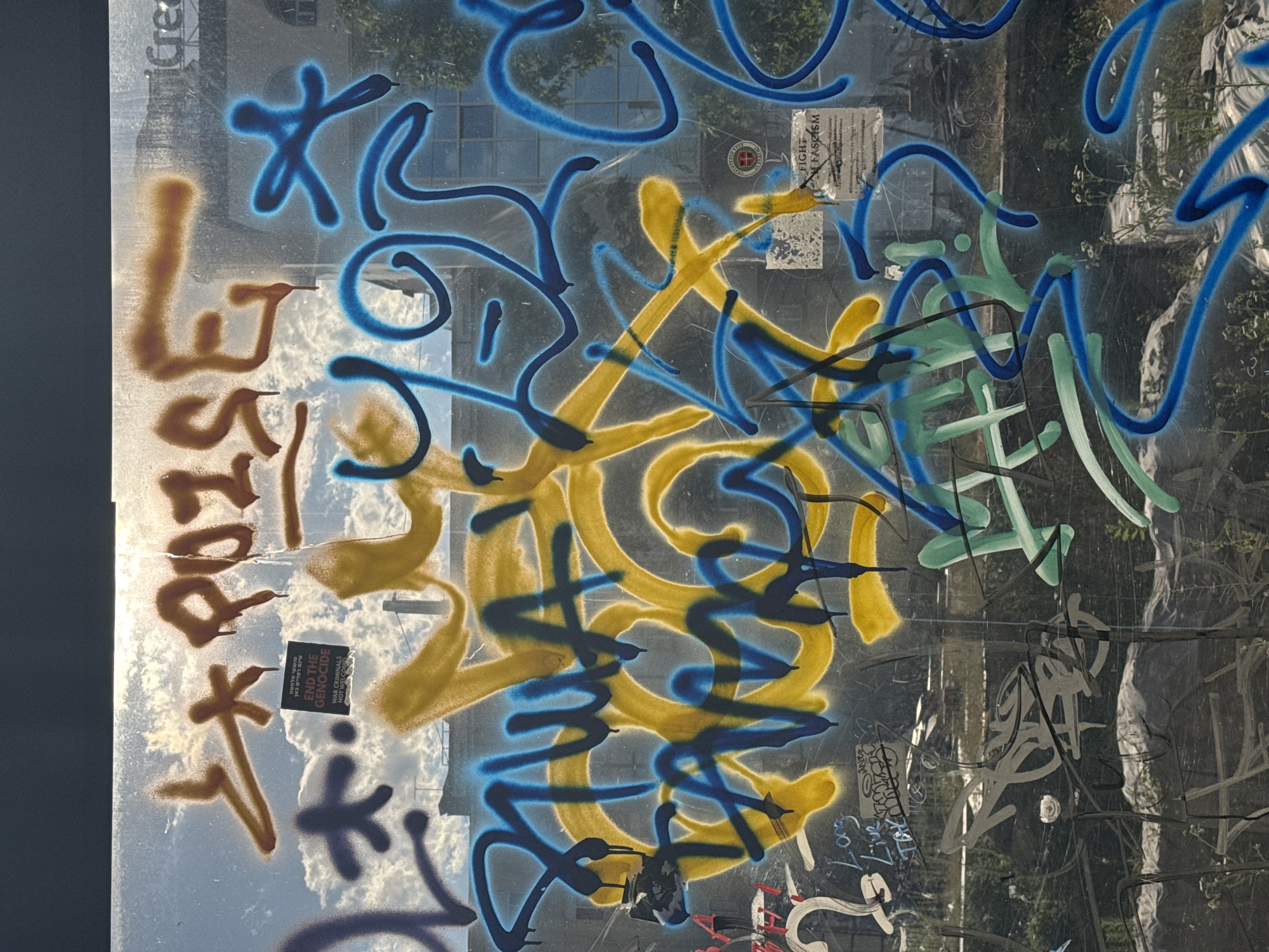 The image captures a close-up view of graffiti on a glass surface, likely a window or storefront, with a blurred urban backdrop featuring buildings and a cloudy sky. The graffiti is predominantly in yellow, blue, and brown spray paint, with some black markings. Prominently displayed on the glass is a sticker with the text "END THE GENOCIDE" and "WAR CRIMINALS NOT WELCOME" in English, and Hebrew script above it. Another sticker near the bottom right corner reads "FIGHT ON FASCISM." The graffiti itself consists of abstract tags and lettering, some dripping paint, obscuring much of the view behind the glass. The overall scene suggests an urban environment with a strong political or social message conveyed through graffiti and stickers. The specific location in the city of Sofia, Bulgaria, is not identifiable from the visual information provided.