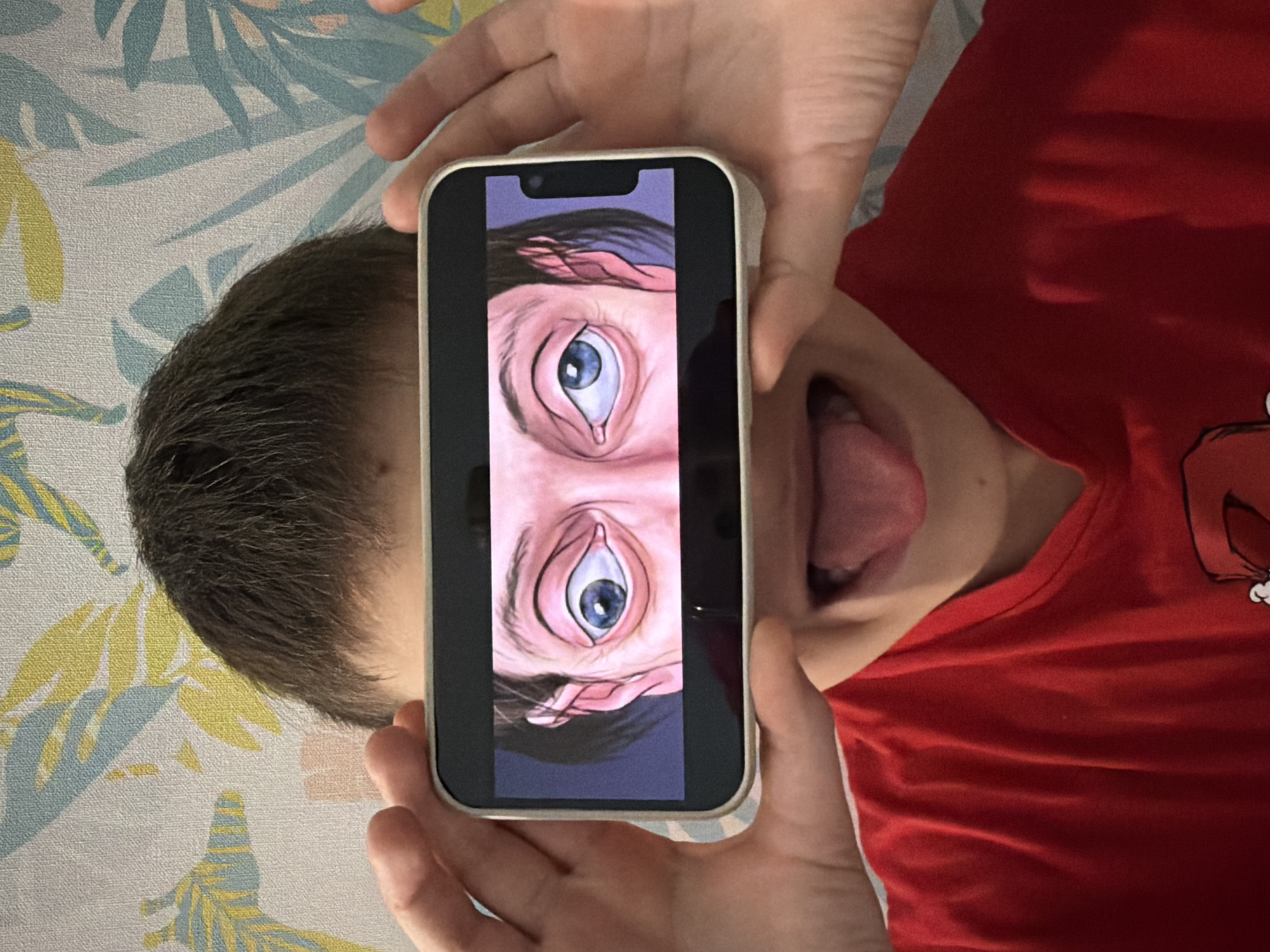 An overhead shot captures a person, possibly a child or young adult, lying down in an indoor setting in Votkinsk, Russia. The individual is wearing a red t-shirt and has dark hair, visible on the left side of the frame. With both hands, they hold a light-colored smartphone vertically directly in front of their face. The person's mouth is wide open with their tongue sticking out, conveying a playful or humorous expression.
The smartphone screen displays a digital artwork featuring two stylized, wide-open eyes with blue irises, seemingly looking upwards, which creates a whimsical visual. The background is a light-colored surface, likely a bedsheet or wallpaper, decorated with a delicate botanical pattern of light blue and yellow leaves. The overall scene suggests a candid and lighthearted moment, with no specific time of day or other location cues beyond the indoor setting. No visible text is present in the image.