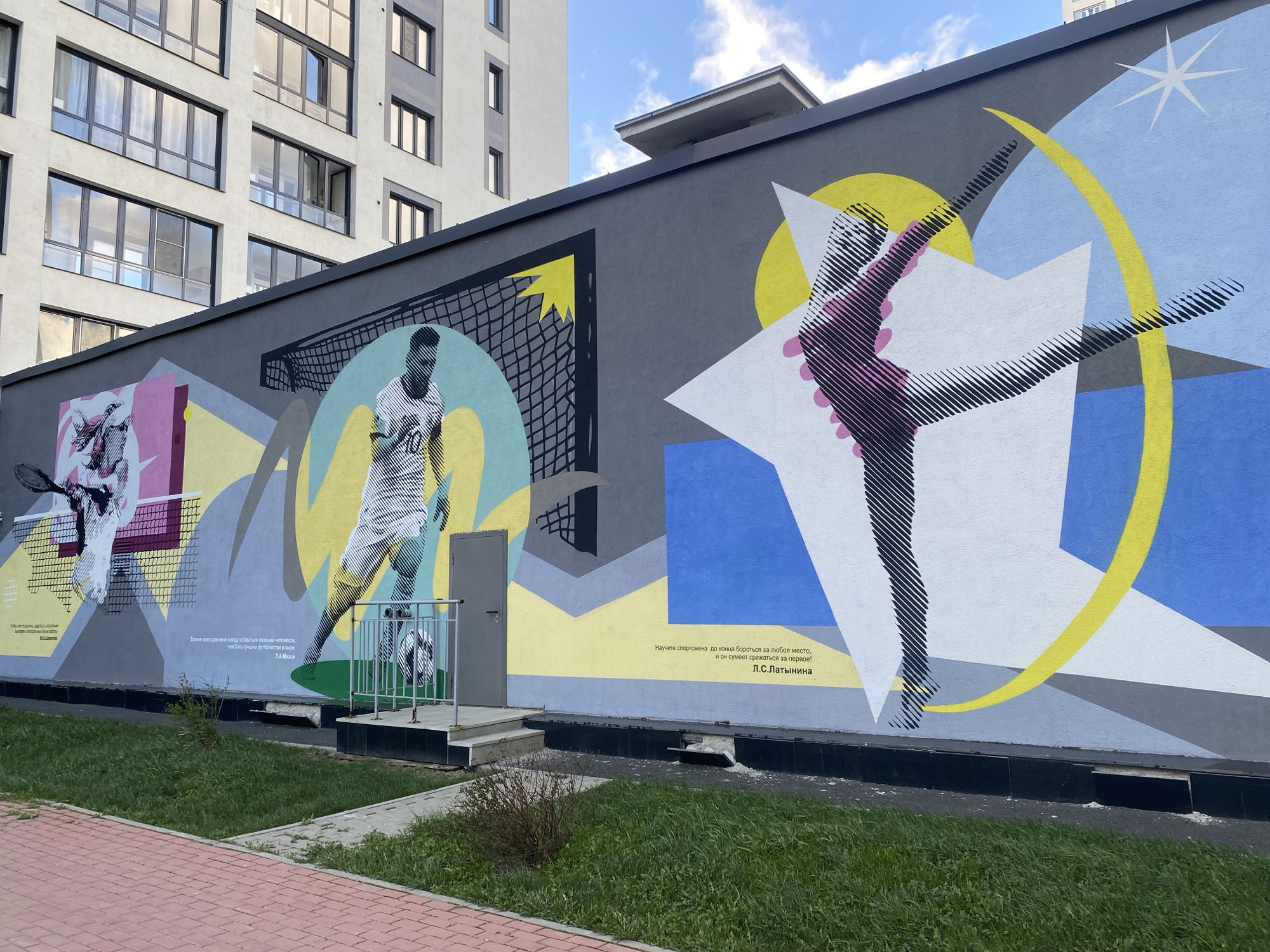 The image depicts a large mural painted on the side of a building in Yekaterinburg, Russia. The mural features representations of three different sports: tennis, soccer, and gymnastics. The tennis portion shows a player with a racket near a net; the soccer portion shows a player kicking a ball, with a goal in the background; and the gymnastics portion shows a gymnast in a mid-air pose, with geometric shapes and a star. Beneath each sport's representation are quotes in Russian. There is a doorway and a small grassy area with shrubs in front of the mural. To the left is a modern apartment building with several windows. The sky is blue with few clouds. A brick walkway is visible on the lower left.