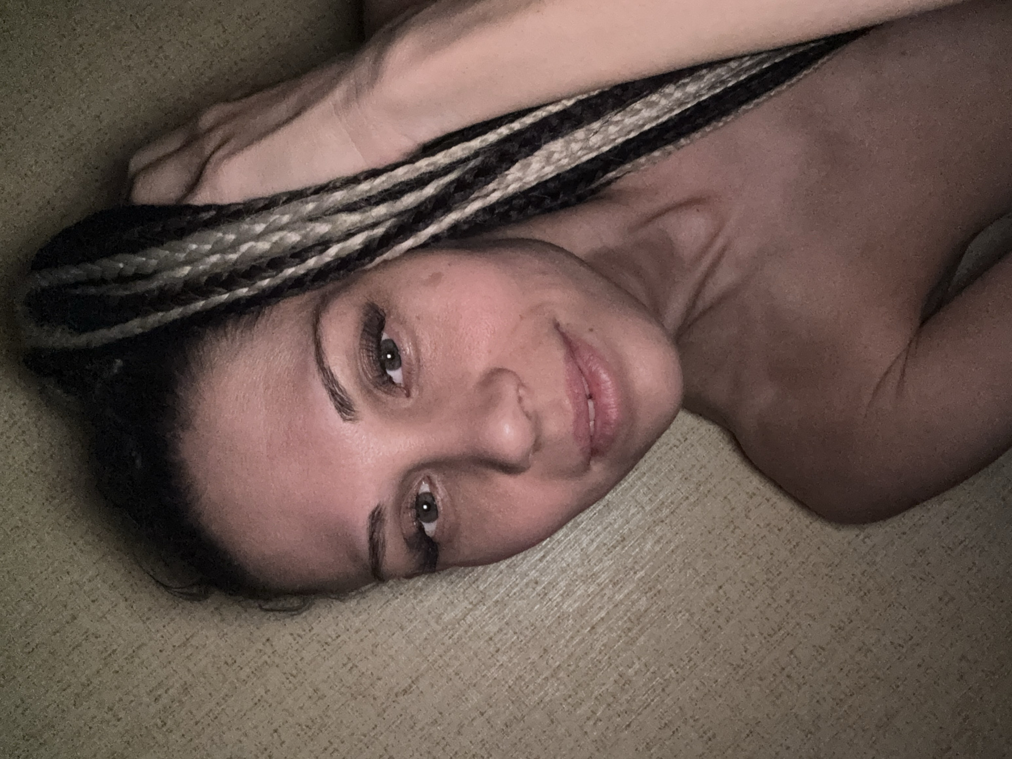 A woman with dark and light braided hair is lying on a textured surface. Her head is tilted towards the camera, and her arm is raised above her head. Her eyes are open, and she has a slight smile. The background is out of focus, showing the texture of the surface she is lying on. The location cannot be determined from the image, and there is no mention of Rusanovo, Russia, in the visible details.