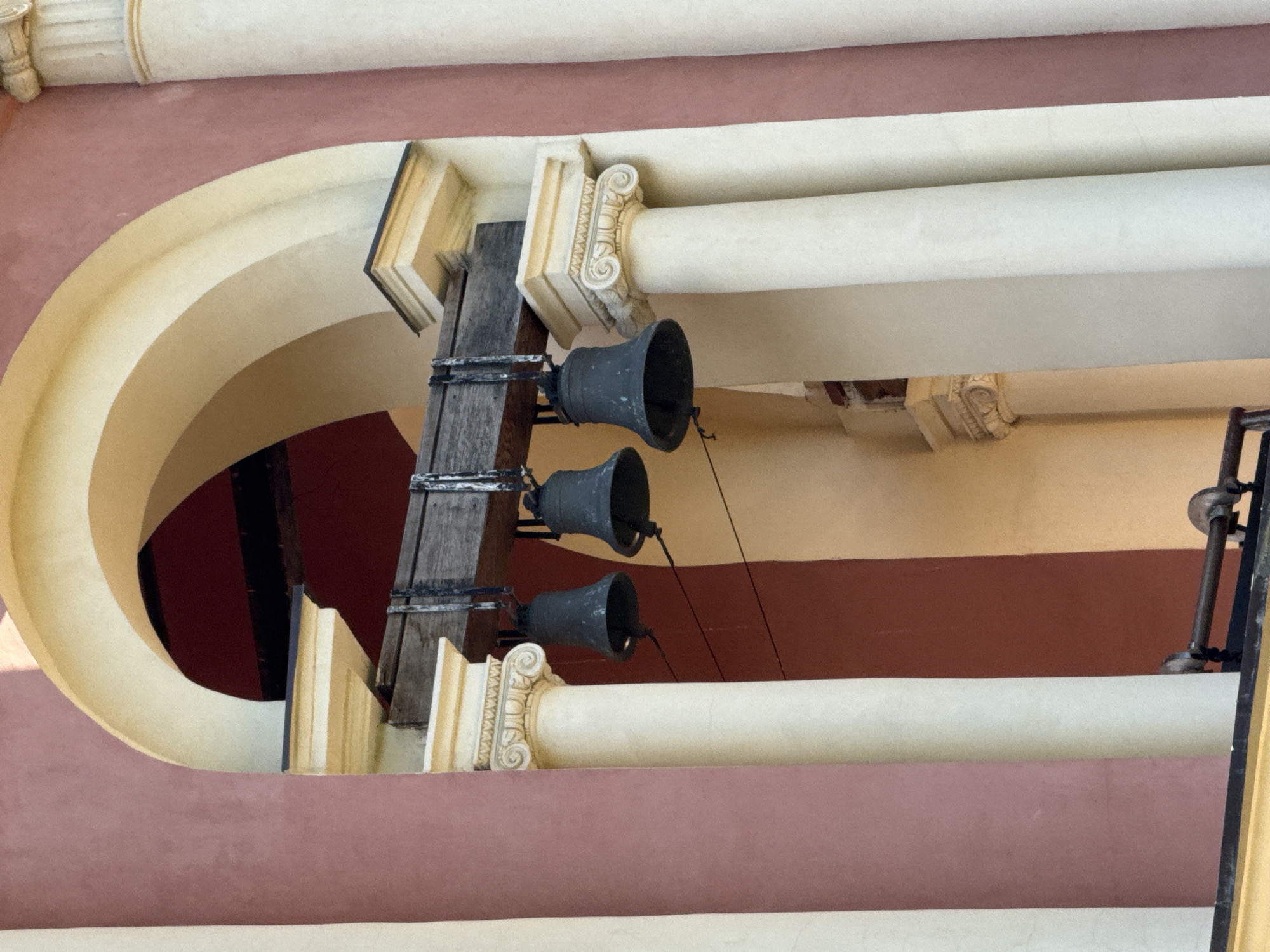 The image shows a close-up view of bells hanging from a wooden beam within an architectural structure. There are three dark-colored bells visible, each suspended by a rope or wire. The bells are of a traditional bell shape and appear to be made of metal. They are arranged vertically on the wooden beam, with two bells closer together and one positioned slightly above and to the side.
The architectural elements surrounding the bells include smooth, curved surfaces in cream and pale yellow, suggesting an archway or decorative recess. There are also columns or pilasters with decorative capitals in a classical style, contributing to the ornate nature of the setting. The background features bands of color, with a darker reddish-brown and a lighter beige or cream, possibly representing different sections of a building's facade or interior. A dark wooden beam, possibly part of a bell tower or belfry structure, holds the bells.
The overall impression is of an ecclesiastical or historical building, likely a church or cathedral, given the presence of bells and classical architectural details. The lighting suggests daytime, with soft shadows indicating ambient light rather than direct sunlight. There is no visible text in the image. The focus is entirely on the bells and their immediate surroundings, creating a sense of stillness and quiet contemplation, despite the potential for the bells to produce sound. The context of Moscow, Russia, reinforces the likelihood of this being a religious building of historical significance.