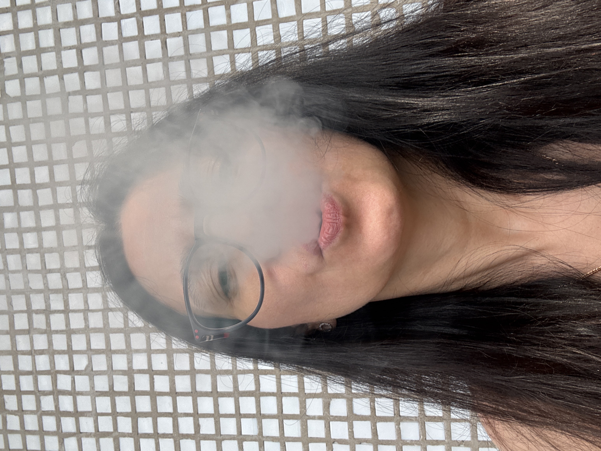 A person is lying down against a white-tiled surface. They are emitting smoke from their mouth, obscuring part of their face. They wear glasses with thin, black frames. The person has long, dark hair that sprawls around their head. A small earring is visible in their ear. The tiled surface has a grid pattern. The location is not discernible from the image.
