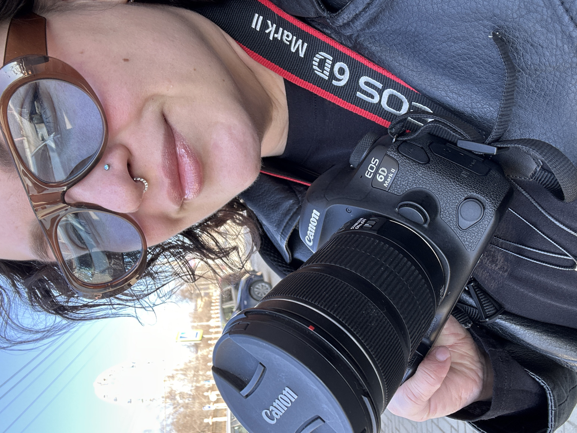 A person takes a selfie, holding a Canon EOS 6D Mark II camera. The camera has a lens with a Canon lens cap. The camera strap says "EOS 6D Mark II". The person wears large, brown sunglasses and has a nose piercing. The reflection in the sunglasses shows the cityscape of Postol, Russia, featuring a Ferris wheel. The person wears a black leather jacket. A car and street signs are visible in the background.