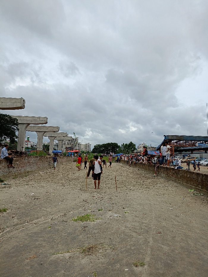 The image depicts an outdoor scene in Sujabad, Bangladesh, under a heavily overcast, grey sky. The central foreground and midground consist of a wide, dry, light-brown earth area, sparsely covered with small green vegetation patches, consistent with a dried riverbed or canal.
To the left, a series of five unfinished concrete support structures, characteristic of an elevated road or bridge construction, extend into the mid-distance. Beyond these structures, an urban landscape featuring multi-story buildings, including a mosque with a green dome and white minarets, is visible. One male in a light blue shirt is seated on a low concrete wall bordering the left side of the dry ground.
In the central dry area, approximately 10-15 individuals, predominantly male and casually dressed, are present. Two vertical wooden sticks are planted in the ground in the foreground, and one male in a white vest and dark shorts holds a long wooden stick, suggesting engagement in a casual game, possibly cricket. Other individuals are distributed across this area, walking or observing.
On the right side, a dark-colored, low-lying wall or embankment borders the dry ground. Approximately 20-30 individuals, predominantly male, are gathered on or near this wall, observing the activity in the central area. Beyond them, informal structures with blue tarpaulins, parked vehicles (including cars and auto-rickshaws), and further urban development are visible in the background.