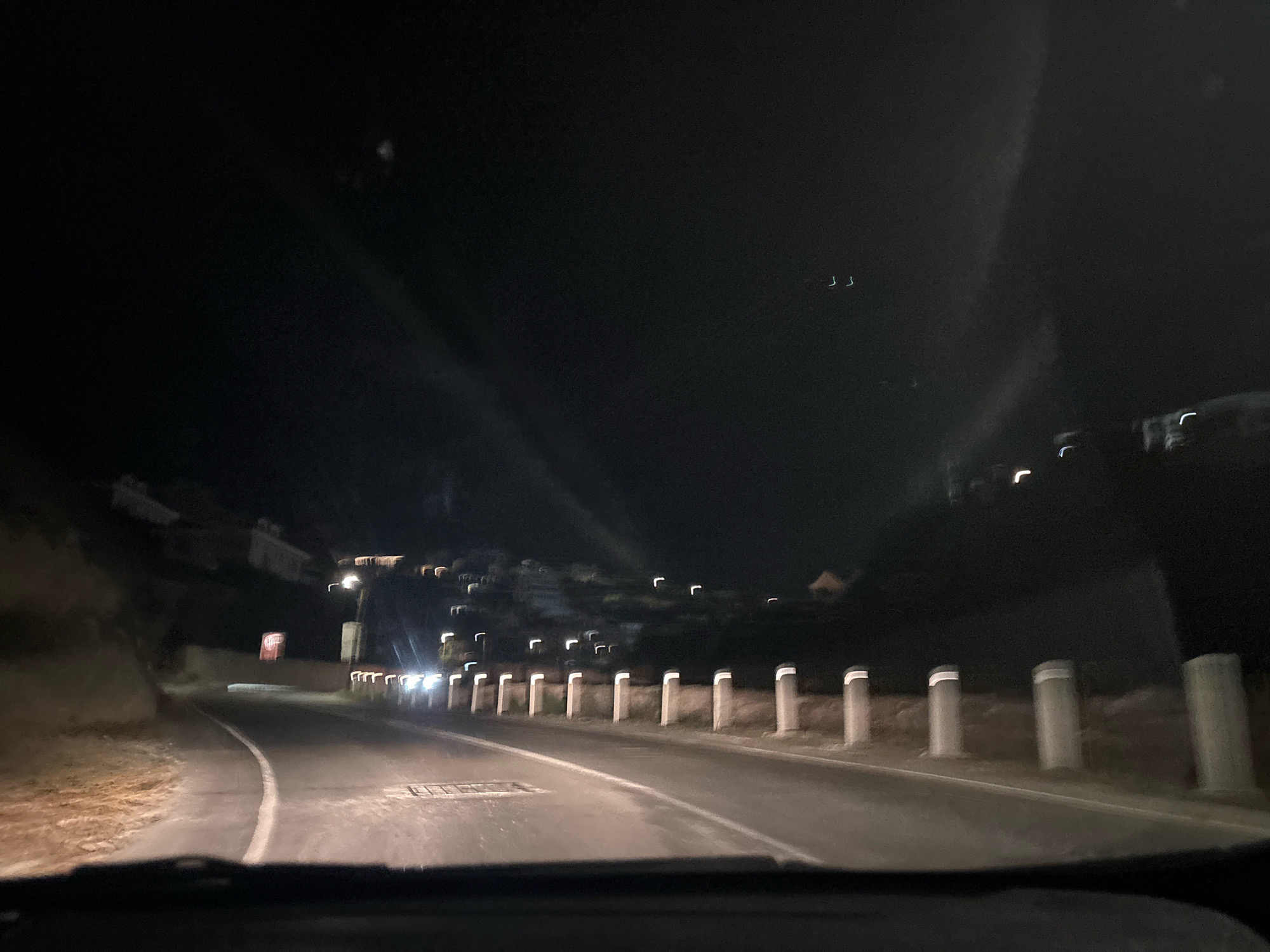 The image depicts a nighttime view of a road in Agios Tychonas Community, Cyprus. The road curves to the left, bordered by a rocky embankment on the left and a series of cylindrical bollards with reflective white bands on the right. Some of these bollards are illuminated by headlights from an approaching vehicle, which also casts light onto the road surface. In the distance, lights from buildings can be seen scattered on a hillside. The sky is dark with some faint, elongated light streaks or reflections visible. There are no discernible people or specific actions captured in the scene.