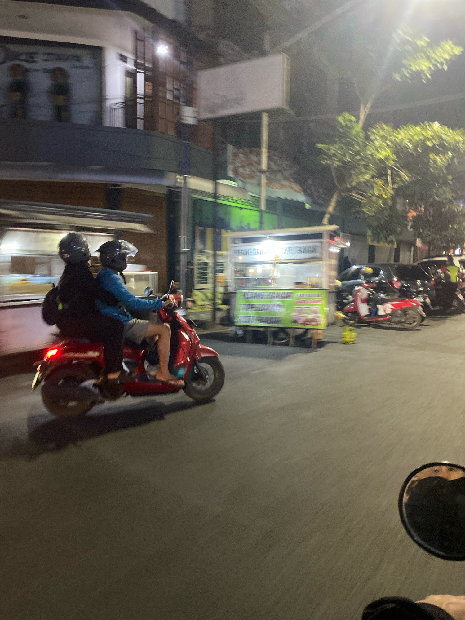 Two individuals wearing helmets are riding on a red scooter at night on a paved street in Kediri City, Indonesia. The person in front is driving, wearing a blue shirt and gray shorts, while the person behind is wearing a black jacket. The scooter is in motion, indicated by the motion blur.
In the background, there are buildings with illuminated windows and signs, and a food stall with bright lighting. Trees are visible, and other vehicles, including motorcycles and cars, are parked or moving in the distance. The scene appears to be a street in a commercial or residential area of the city at night, with streetlights and light emanating from the buildings and stalls. The overall impression is a typical urban night scene in Indonesia.