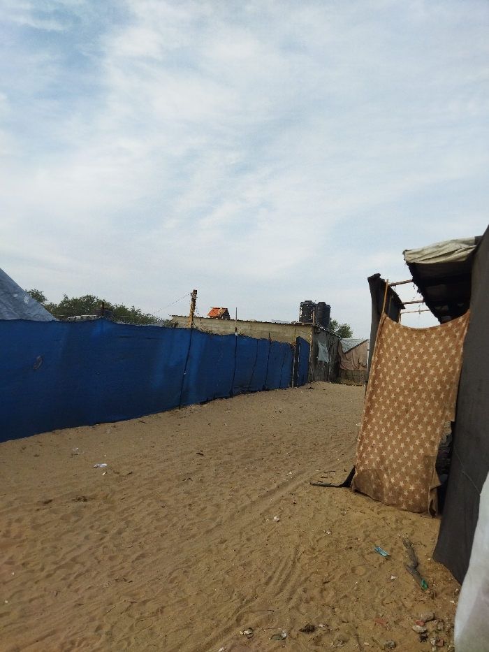 A sandy pathway runs between makeshift dwellings. On the left, a long blue fence stands, topped with remnants of buildings. To the right, a draped curtain with star patterns hangs from a simple structure. Litter is scattered on the sandy ground. The sky is overcast with sparse clouds. A water tank sits atop a building further down the path. The scene depicts a poverty-stricken area.