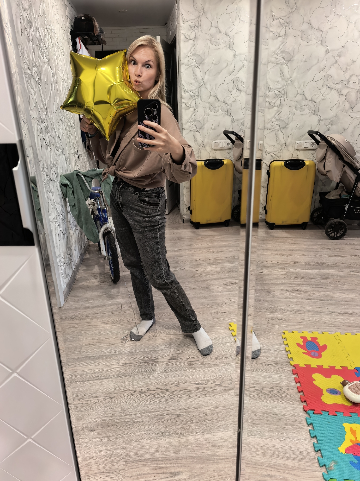 An adult female is depicted in a full-length mirror, capturing a selfie in what appears to be a residential hallway or entryway in Sterlitamak, Russia. The woman, who has shoulder-length blonde hair styled straight, is making a playful, exaggerated facial expression with puckered lips and wide eyes. She is wearing a light brown or taupe long-sleeved shirt, tied in a knot at the waist, and dark grey faded jeans. Her feet are clad in white ankle socks with grey heels and toes. In her left hand (viewer's left), she holds a shiny gold star-shaped Mylar balloon by its string. In her right hand, she holds a black smartphone with a dark patterned case, visible due to the mirror reflection as she takes the photograph.
The background, visible through the mirror, features walls with a distinctive white finish adorned with intricate grey or silver marbled patterns. The floor is covered with light wood-look laminate or vinyl plank flooring in greyish-brown tones.
To the left of the woman's reflection, behind a large white door frame or wall section, a small blue child's bicycle with white tires is parked against the patterned wall. A green cloth or jacket is draped over its handlebars. Above the bicycle, obscured by the woman's head, are dark objects on a shelf or rack, possibly bags or clothing items.
To the right of the woman's reflection, two identical bright yellow hard-shell suitcases with black accents (handles, wheels) are visible, standing upright against the wall. Above them are two white wall plates with electrical outlets. Next to the suitcases, a beige or tan baby stroller, folded or partially collapsed, with a black frame and the "WONDERKID" brand name visible, is parked. On the floor in the foreground on the right, a multi-colored children's interlocking foam playmat (yellow, red, blue, turquoise) with cut-out animal shapes is laid out. A small white or beige electronic device, possibly a remote or toy, rests on the playmat. A small yellow object, likely a piece of a toy or the playmat, is near the woman's feet.