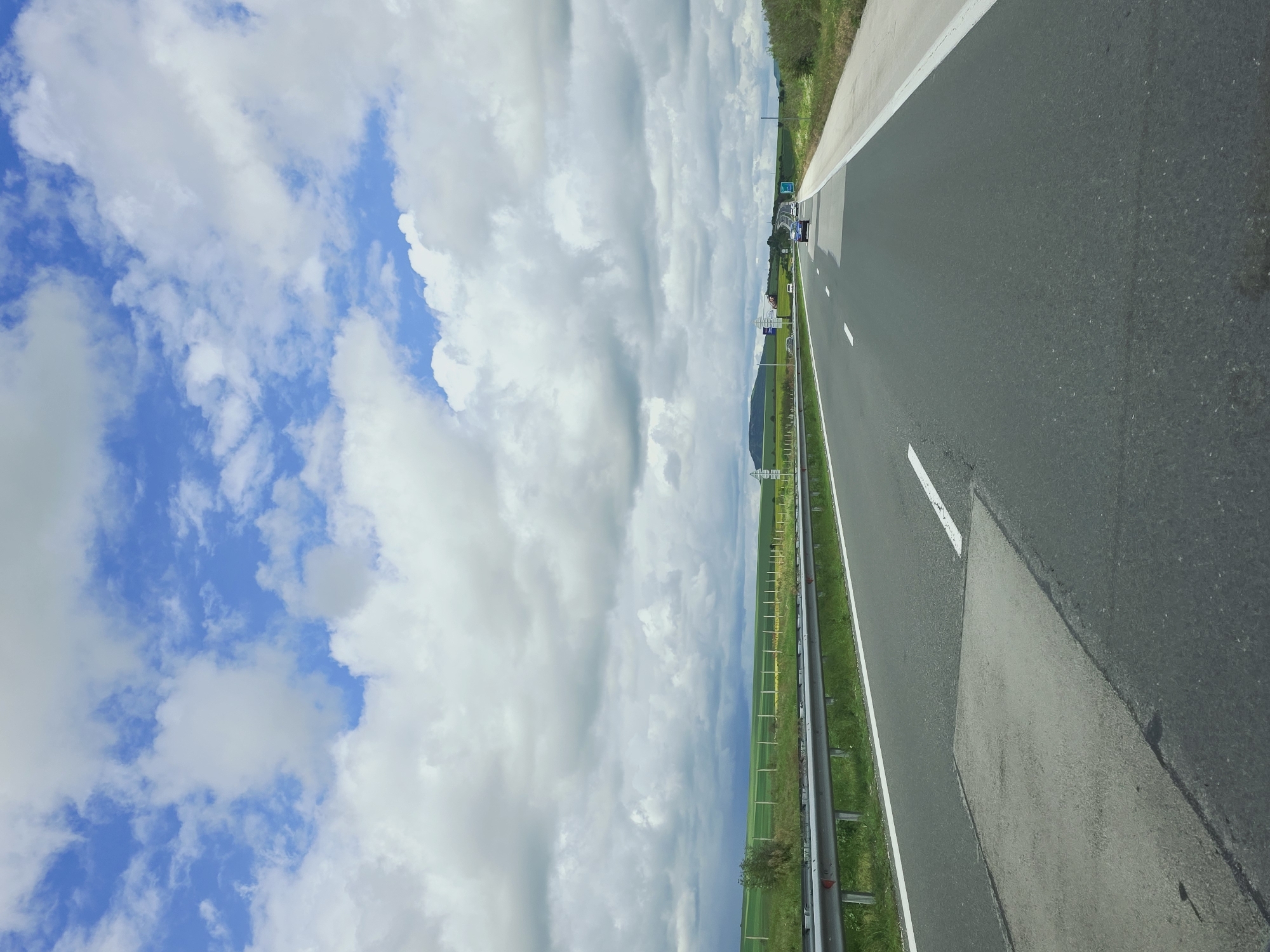A road in Venchan, Bulgaria, is shown from a vehicle's perspective. The road, with white lines, is asphalt and has a concrete barrier on the right side. To the left of the barrier is a grassy verge. Cars are visible ahead on the road. The sky to the left is blue with white clouds. A metal barrier is visible. A mountain is in the background.