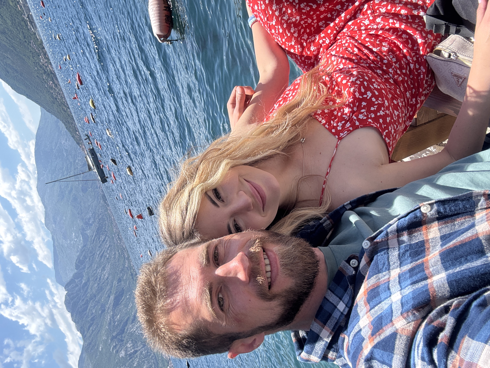 A couple is in a selfie on what appears to be a boat or a dock. The woman, wearing a red dress with white floral patterns, is leaning towards the man, who is wearing a blue and orange plaid shirt over a green undershirt. Behind them is water with some buoys, a sail boat, and mountains visible in the distance. The location is likely Dražin Vrt, Montenegro, given the query.