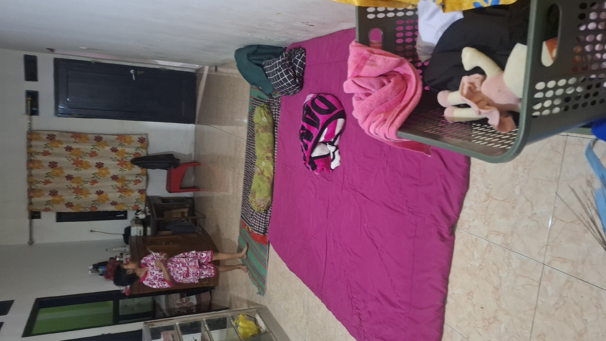 In a room in Cibinong Hilir, Indonesia, a young child wearing a pink and white patterned outfit stands on a tiled floor. To the child's left, a tall wooden cabinet with shelves is visible. In the center of the room, a dark wooden desk with items on its surface is positioned in front of a curtain with a floral pattern. A red stool is placed near the desk. A dark blue door is visible on the right side of the room. A large magenta mat covers a significant portion of the floor. On this mat, a folded blanket with black, pink, and white patterns rests. Further back, on another mat, a green pillow with a floral pattern is situated. To the right of the green pillow, a stack of folded clothes, including a dark green garment and a black and white checkered garment, is present. In the foreground on the right, a dark gray laundry basket contains various items of clothing, including a pink towel and a stuffed toy. The walls are painted white.