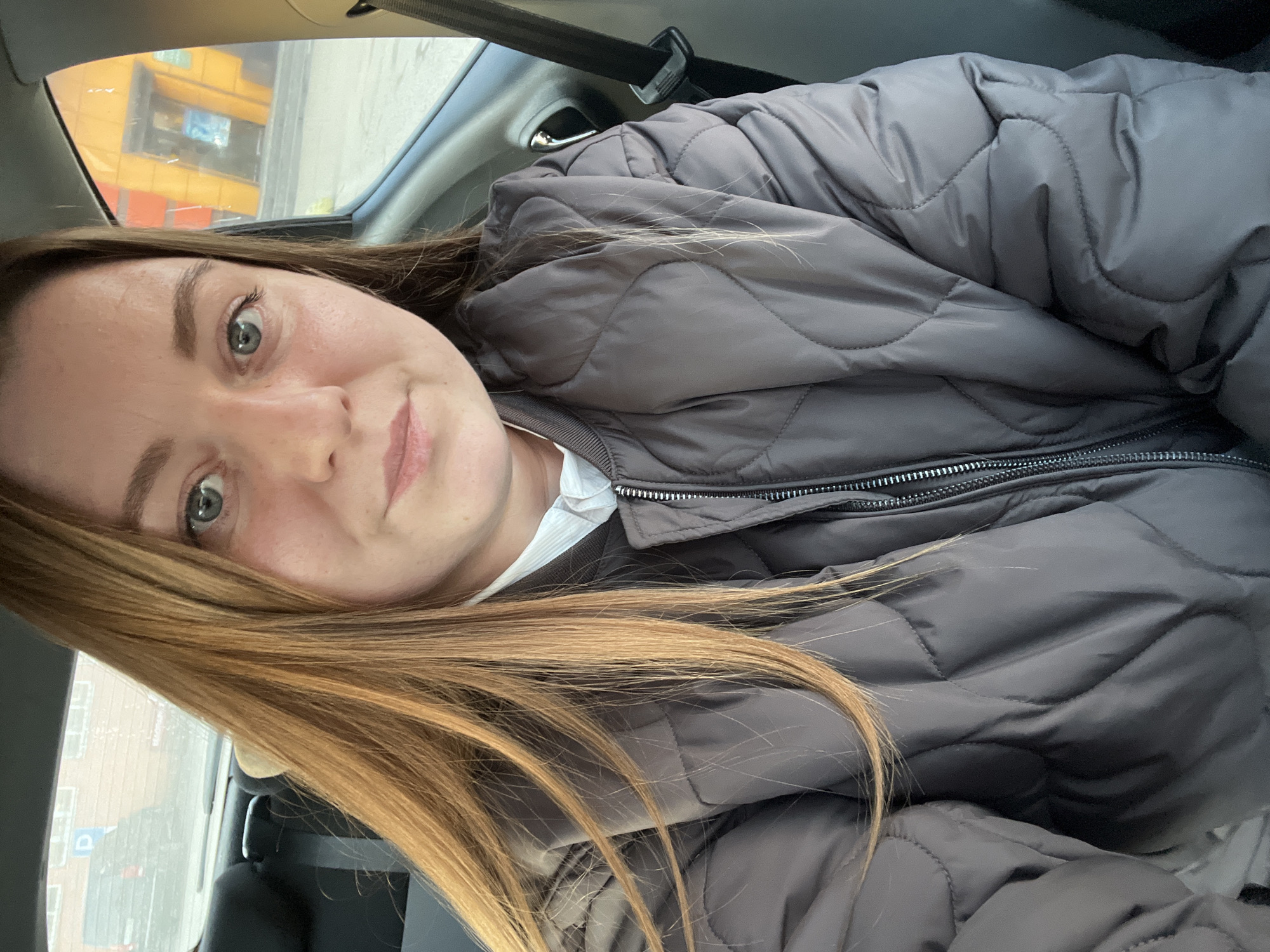 An adult female, with long light brown hair and light blue-green eyes, is seated in the passenger side of a vehicle. She wears a dark grey quilted puffer jacket over a white collared shirt, and is secured by a black seatbelt visible on the right. She looks directly at the camera, implying a selfie.
Through the rear left window, a dark brick building with a white rectangular parking sign (blue 'P' symbol) is visible. Streaks on the window suggest rain or moisture. A dark vehicle is partially obscured behind the brick building.
Through the front right window, a modern commercial building with distinctive orange, yellow, and dark grey paneling is visible. This building has recessed sections, possibly indicating an entrance, with some blue elements, likely signage or advertisements. A sidewalk or pedestrian area is visible in front of the building.
The scene depicts an urban commercial district within Klin, Russia, characterized by modern retail architecture and street infrastructure.