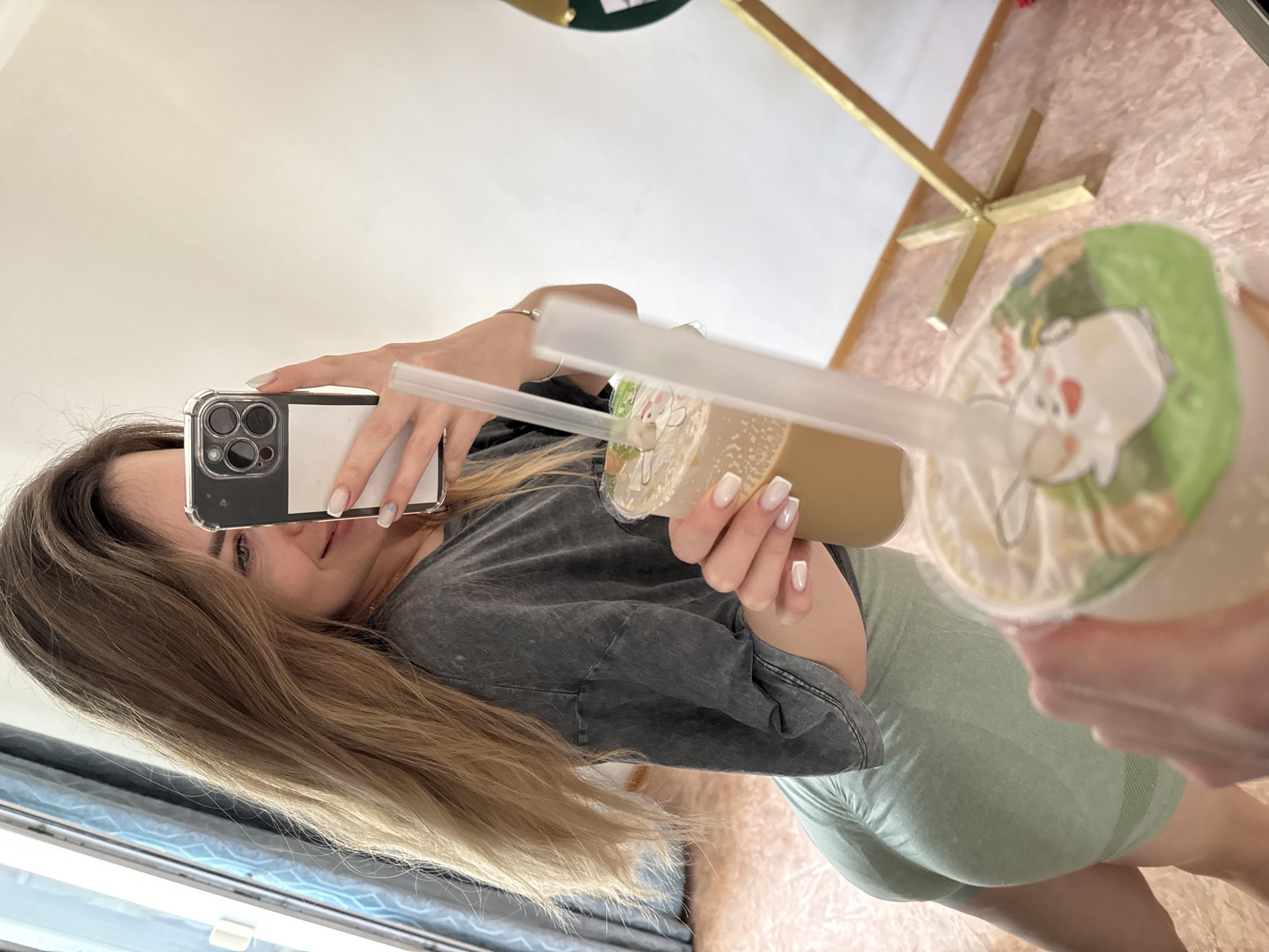 A mirror selfie depicts a young woman holding two plastic cups containing beverages with straws. The woman is partially visible, wearing a grey top and light green leggings, with her long blonde hair cascading to the side. She is holding a smartphone in her left hand, partially obscuring her face, to take the photo. The drinks are in her right hand. A portion of a gold-colored decorative stand is visible in the background, resting on what appears to be a pink, textured surface, possibly a rug. The environment includes a window with blue curtains visible in the lower-left corner of the frame. Location: Unknown, Unknown.