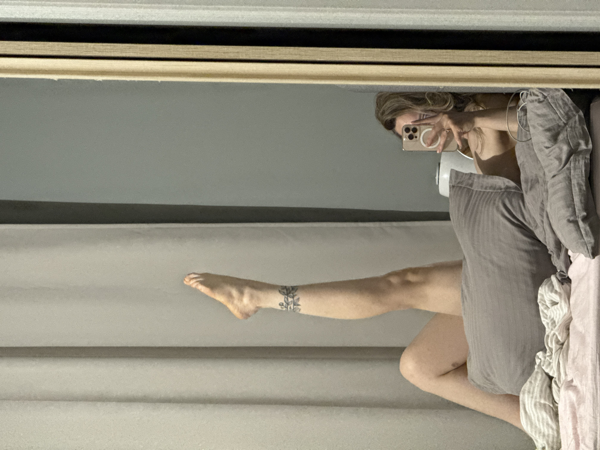 A reflection in a mirror shows a woman in a bedroom, likely in Chaikovsky, Russia, based on location assumptions. She is sitting on a bed with grey and pink bedding, covered with a grey blanket, and holding a smartphone to take a picture of herself. One leg is extended towards the left side of the frame. The woman has light hair and is wearing headphones. A tattoo is visible on her ankle. The background is a grey wall. The mirror has a light-colored frame.