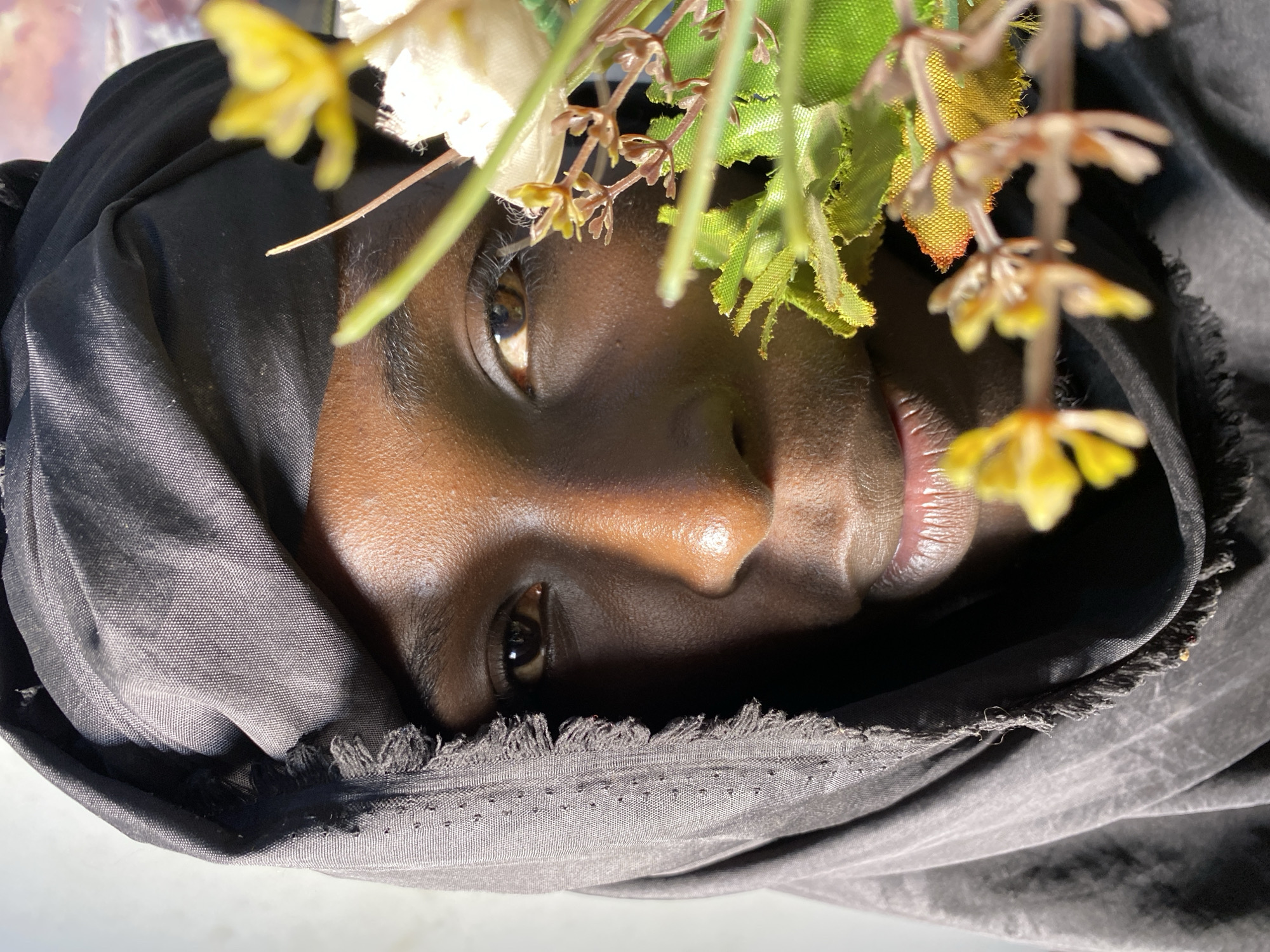 A close-up portrait of a person wearing a black headscarf shows their face partially obscured. The person's right eye, nose, and lips are visible. They have brown skin and are looking towards the viewer. In the foreground, a blurred cluster of artificial yellow and white flowers and green leaves partially covers the right side of the person's face. The background is a blurry mix of light and color, with hints of a sunset sky visible on the right. The image appears to have been taken outdoors during daylight. The person's location in Bukuru, Nigeria is not evident from the visual information provided.