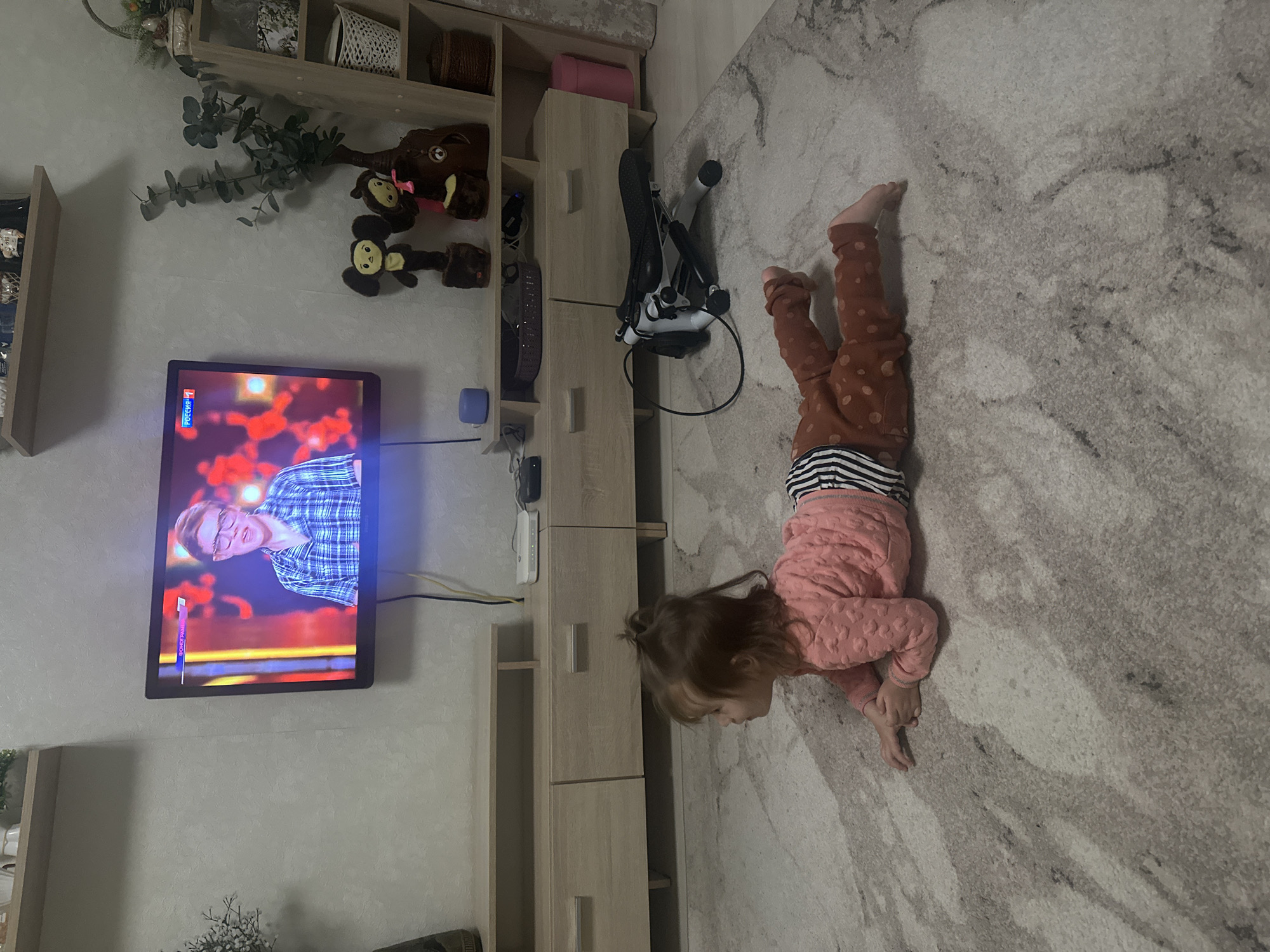 A child is lying on a rug in a room in Uva, Russia. The child is wearing brown pants with polka dots, a striped shirt, and a pink top. The child is lying on their stomach, with their head near a light-colored wooden cabinet. A television is mounted on the wall, displaying a man with glasses on screen. A stroller is parked near the cabinet. A shelf with plants and toys is visible on the wall above the television.