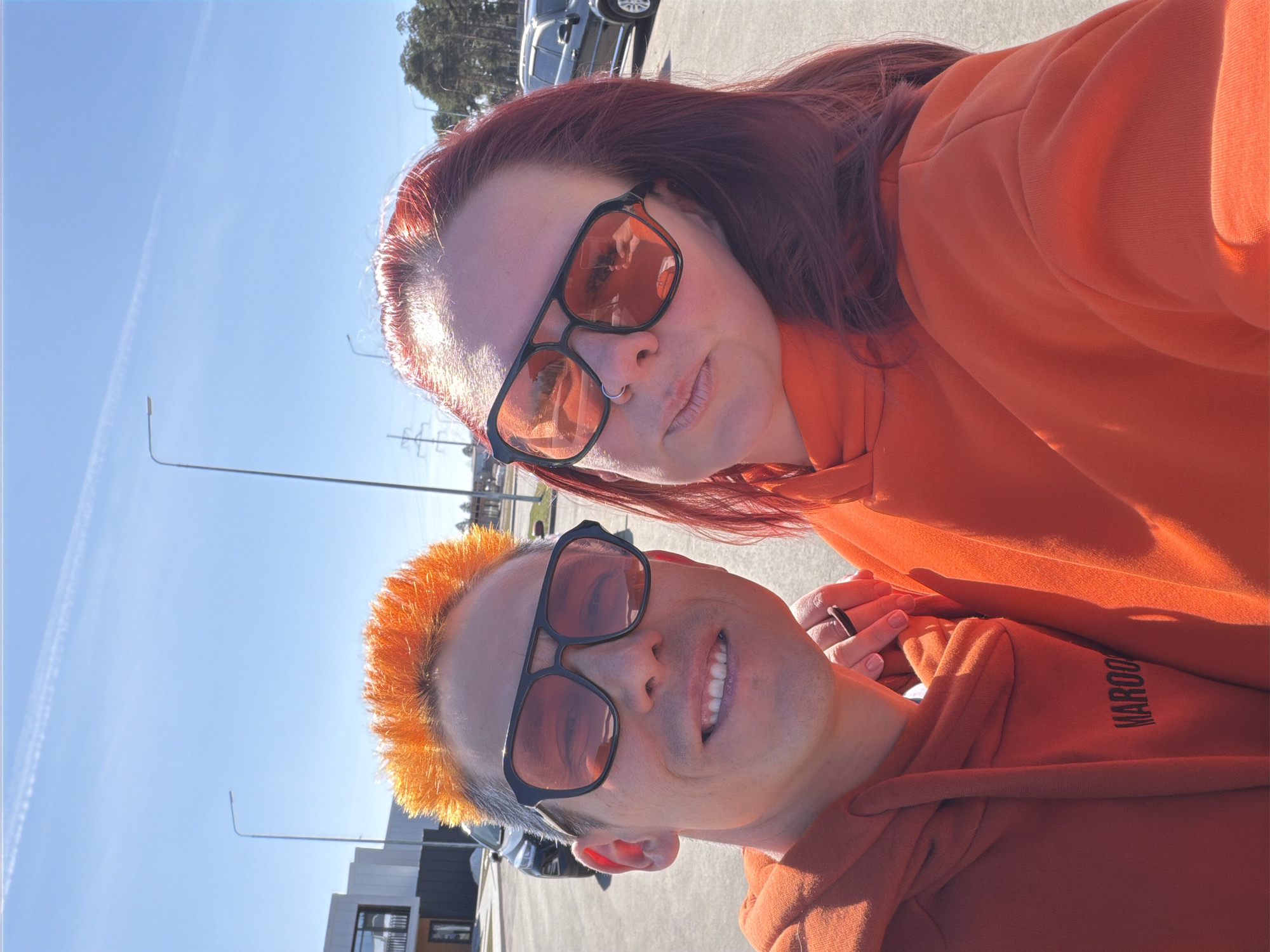 Two individuals are posing for a selfie outdoors. The person in the lower part of the frame has short, bright orange hair and is wearing sunglasses. The person above them has reddish-purple hair and is also wearing sunglasses. Both are wearing similar orange hooded sweatshirts. The sweatshirt of the person in the lower portion of the frame has the word "MAROON" printed on it.
In the background, there is a clear blue sky, streetlights, vehicles, and a modern building with large windows. The location is difficult to ascertain specifically as Votkinsk, Russia, without more distinct markers in the environment.