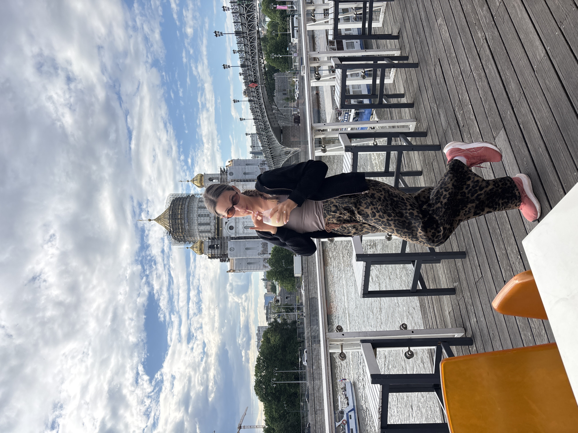 A woman stands on a wooden deck, likely part of a restaurant or tourist area. She wears a black jacket, beige top, leopard-print pants, and rose-colored sneakers and sips from a glass with a straw. The setting is outdoors, under a sky with scattered clouds.
The background features the Cathedral of Christ the Savior in Moscow, Russia, visible on the left. A bridge spans a body of water behind her, adding to the city’s scenery. Tables and chairs suggest an eating area on the deck.